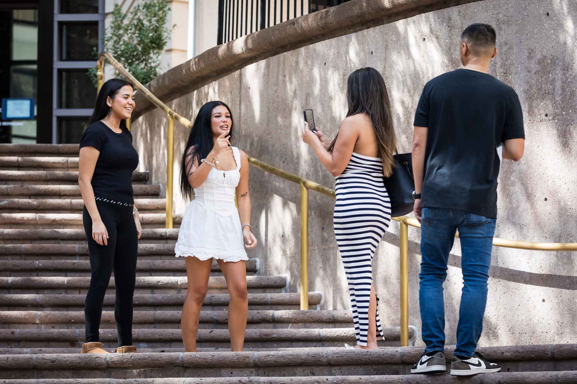 Two young girls and woman wearing striped dress holding phone in front of man wearing jeans on stairs during Riverwalk surprise proposal in San Antonio