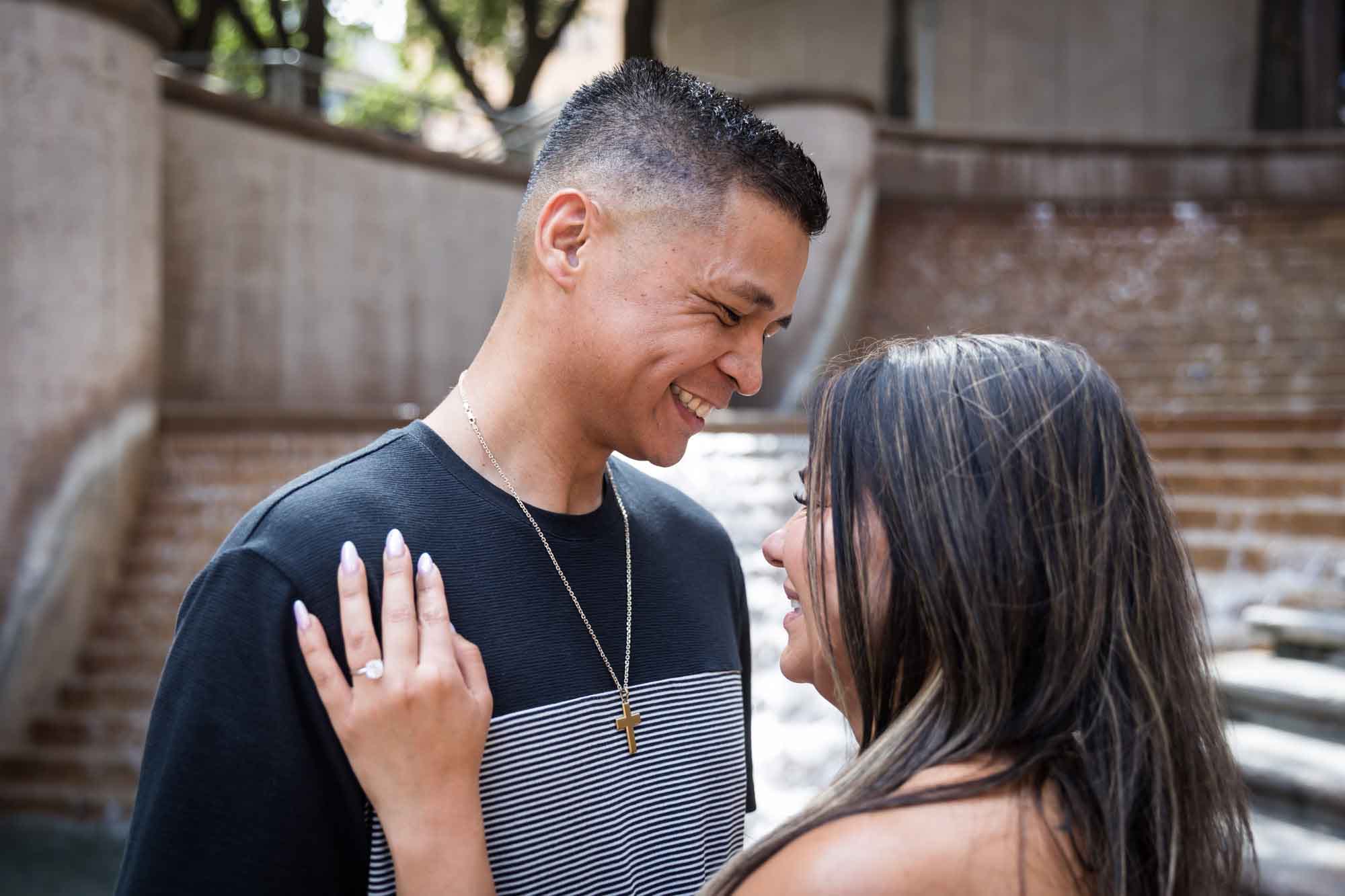 Woman wearing black and white striped, sleeveless dress with hand showing engagement ring resting on man wearing striped shirt during a Riverwalk engagement photo shoot with waterfall and stairs in background