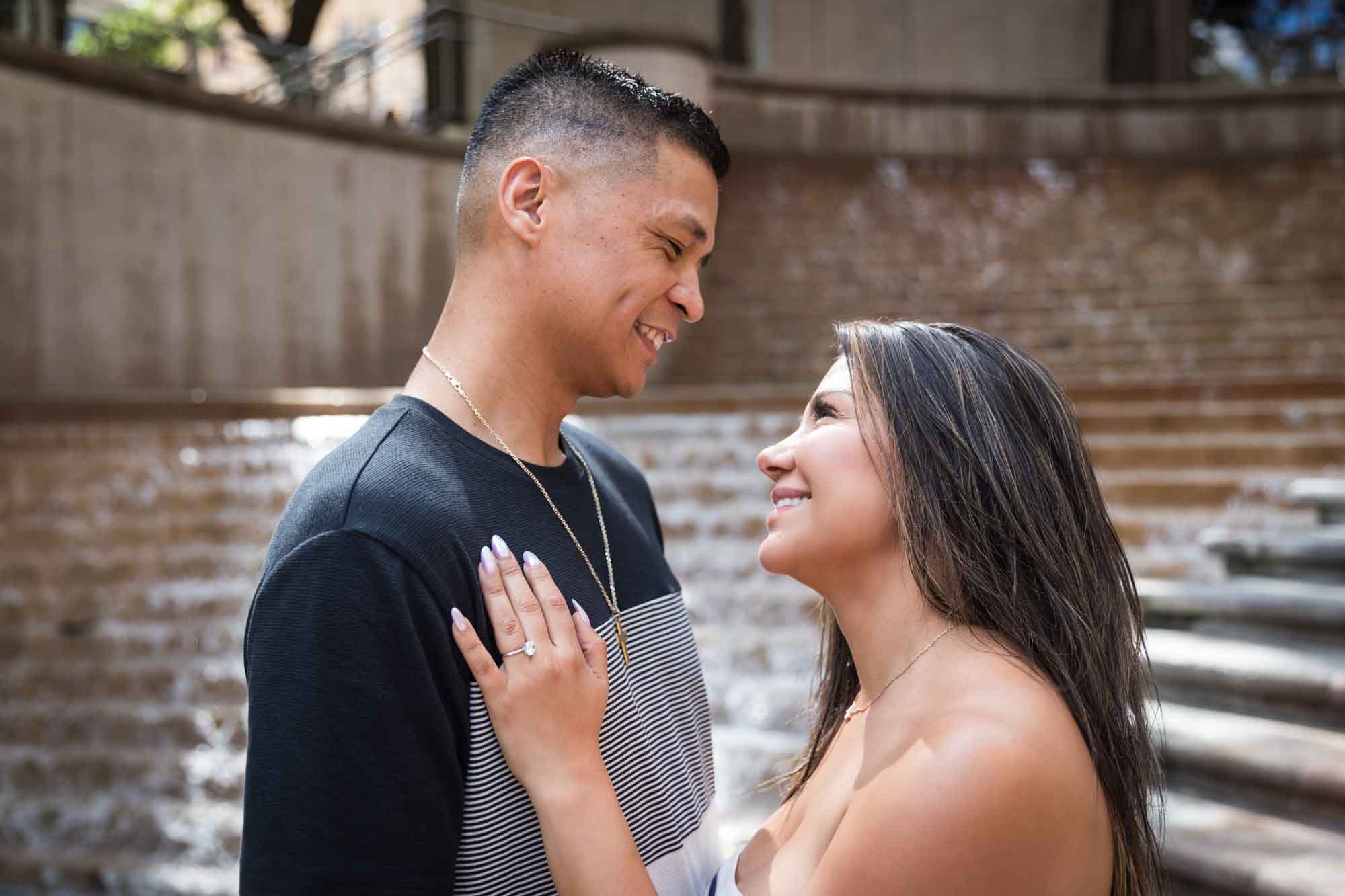 Woman wearing black and white striped, sleeveless dress with hand showing engagement ring resting on man wearing striped shirt during a Riverwalk engagement photo shoot with waterfall and stairs in background