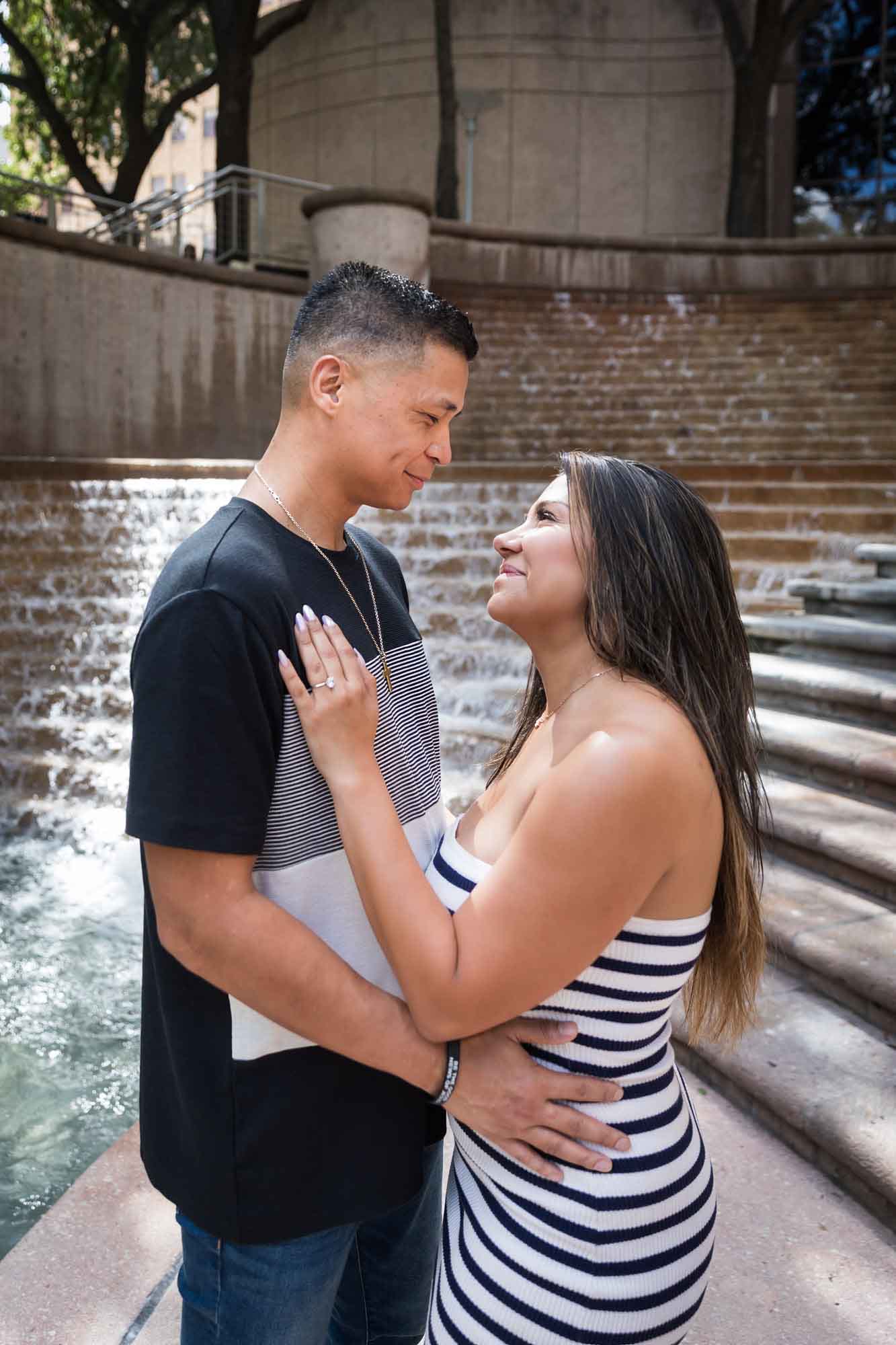 Woman wearing black and white striped, sleeveless dress with hand showing engagement ring resting on man wearing striped shirt during a Riverwalk engagement photo shoot with waterfall and stairs in background