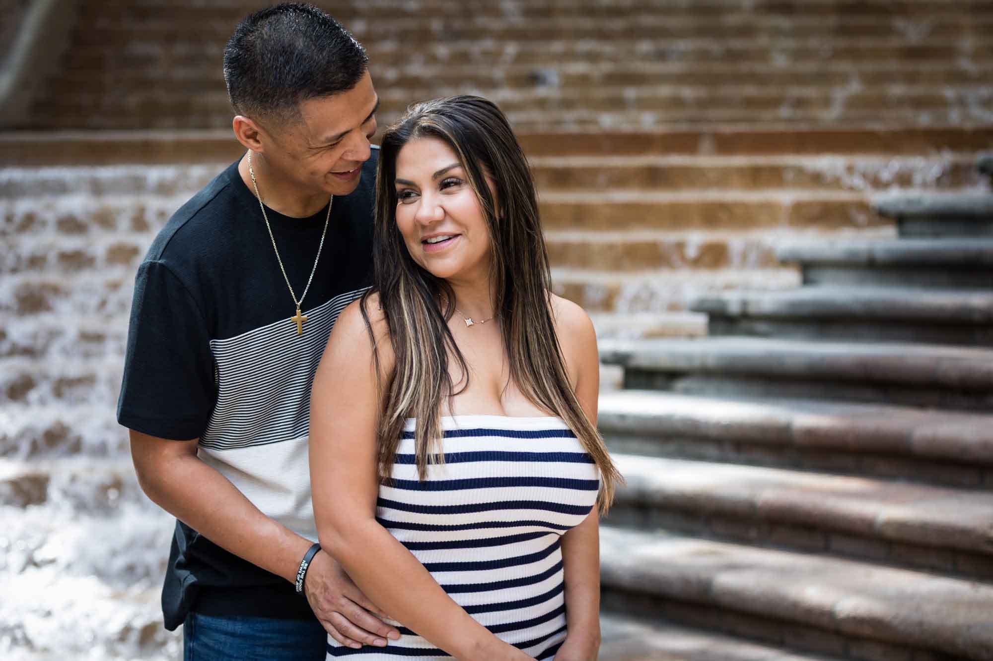 Man wearing striped shirt and jeans hugging woman wearing black and white striped, sleeveless dress during a Riverwalk engagement photo shoot with waterfall and stairs in background