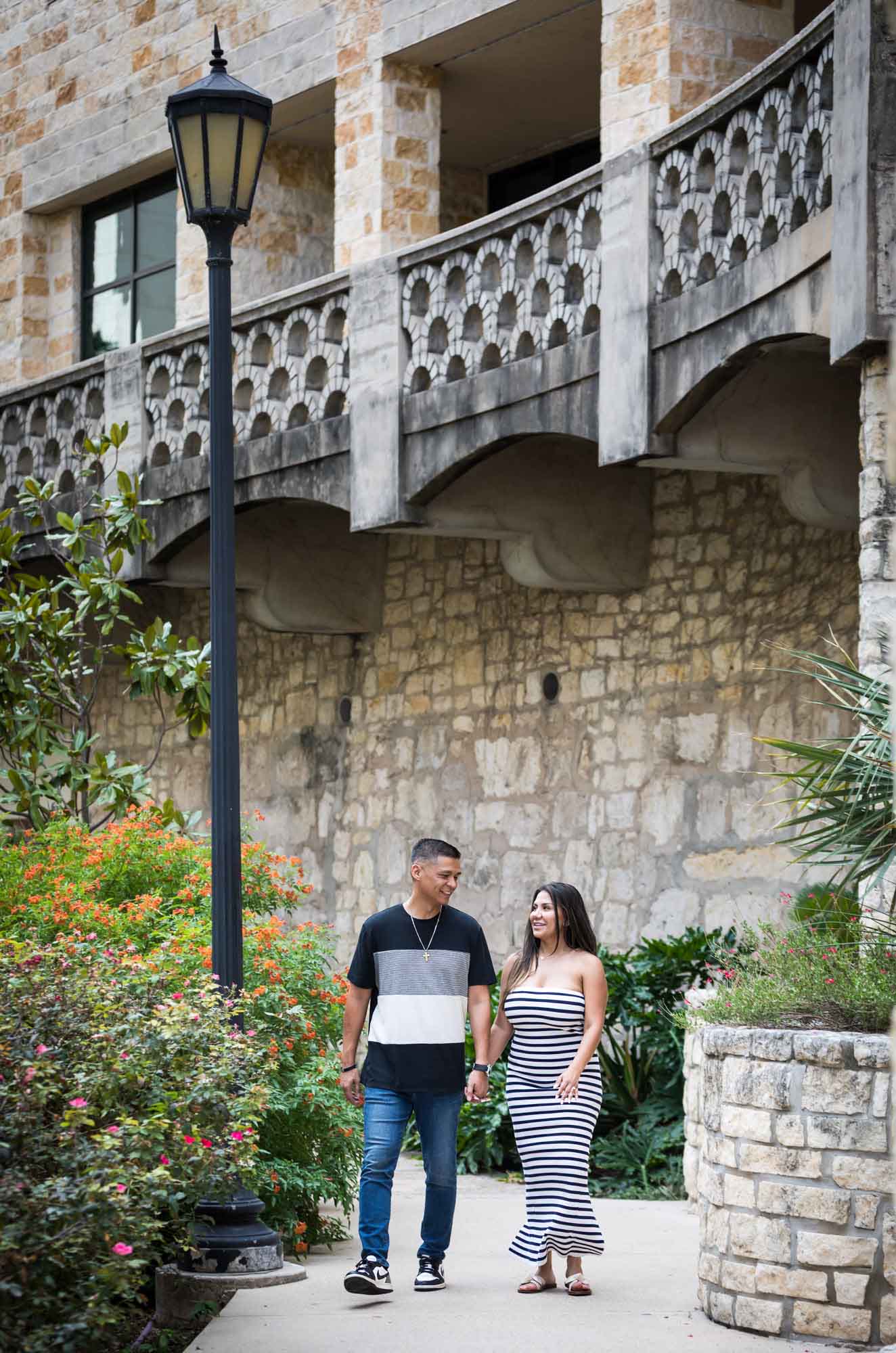 Man wearing striped shirt and jeans holding hands with woman wearing black and white striped, sleeveless dress during a Riverwalk engagement photo shoot with lamp post in the background