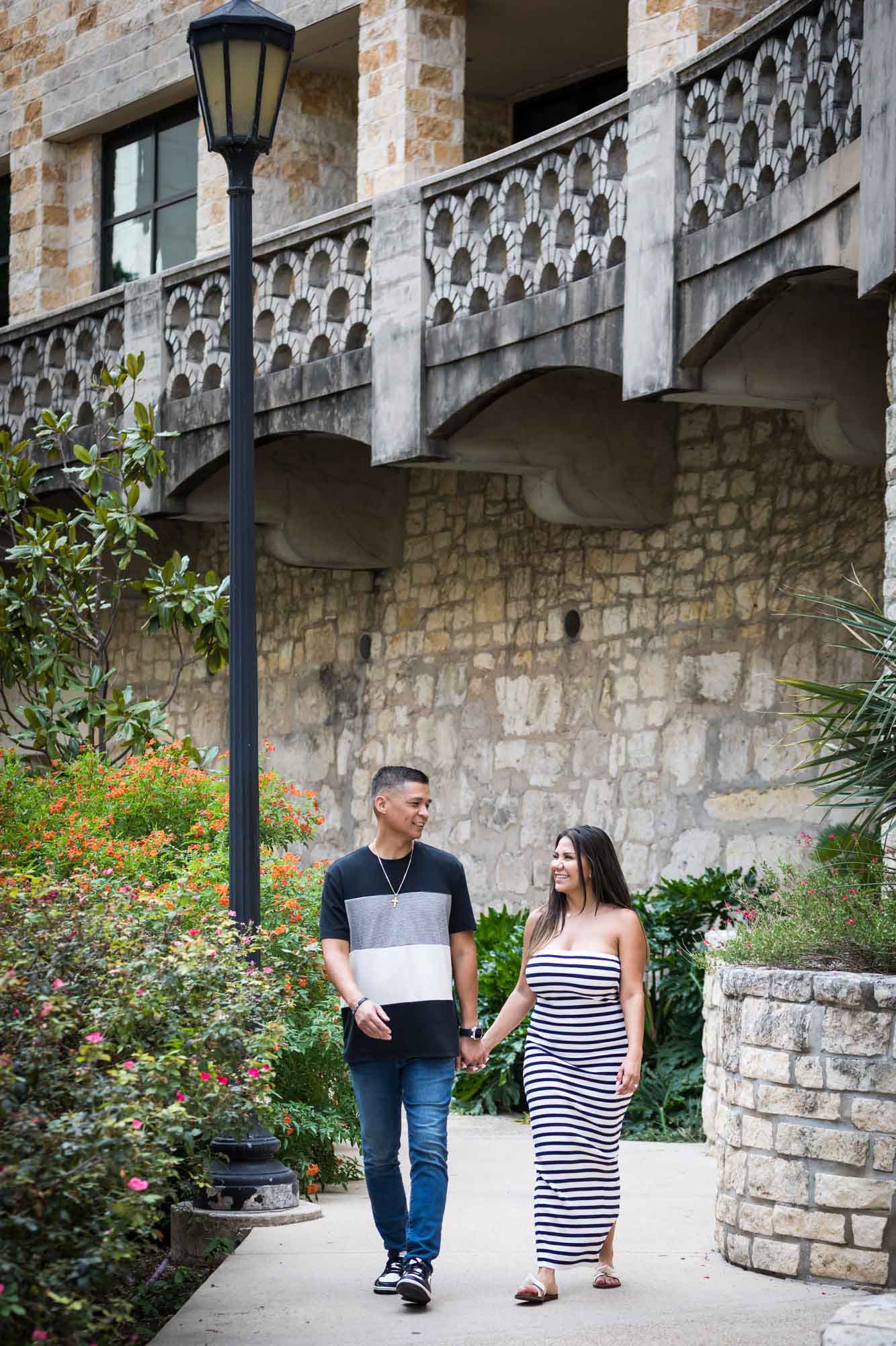 Man wearing striped shirt and jeans holding hands with woman wearing black and white striped, sleeveless dress during a Riverwalk engagement photo shoot with lamp post in the background