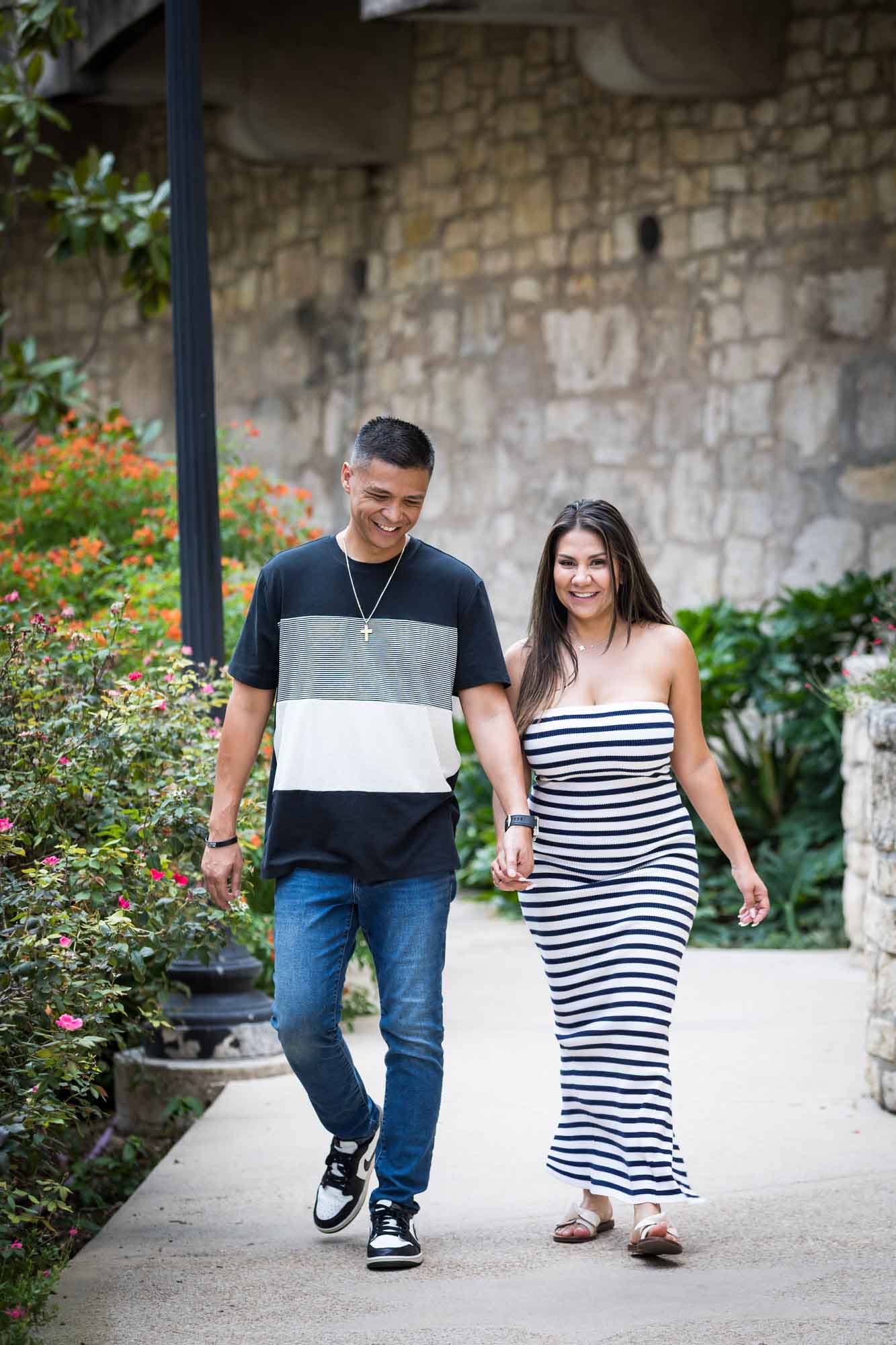 Man wearing striped shirt and jeans holding hands with woman wearing black and white striped, sleeveless dress during a Riverwalk engagement photo shoot