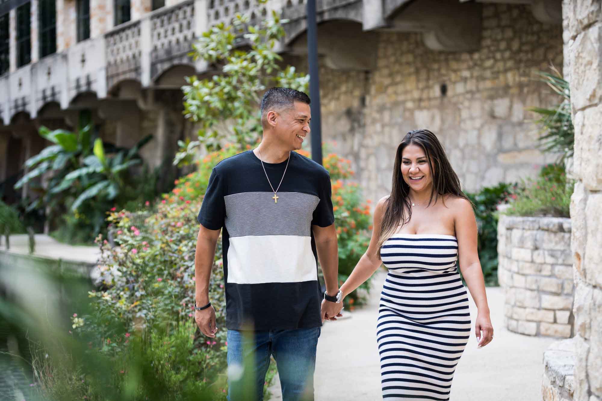 Man wearing striped shirt and jeans holding hands with woman wearing black and white striped, sleeveless dress during a Riverwalk engagement photo shoot