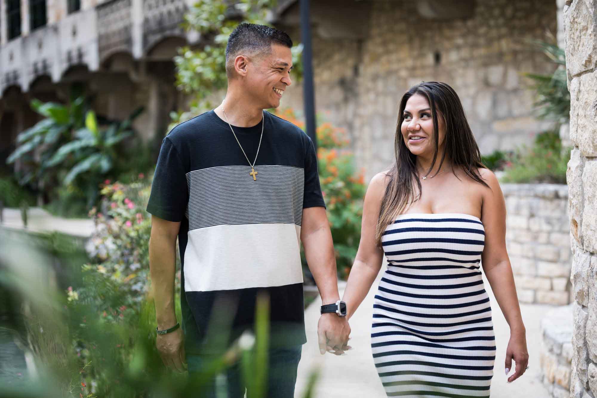 Man wearing striped shirt and jeans holding hands with woman wearing black and white striped, sleeveless dress during a Riverwalk engagement photo shoot