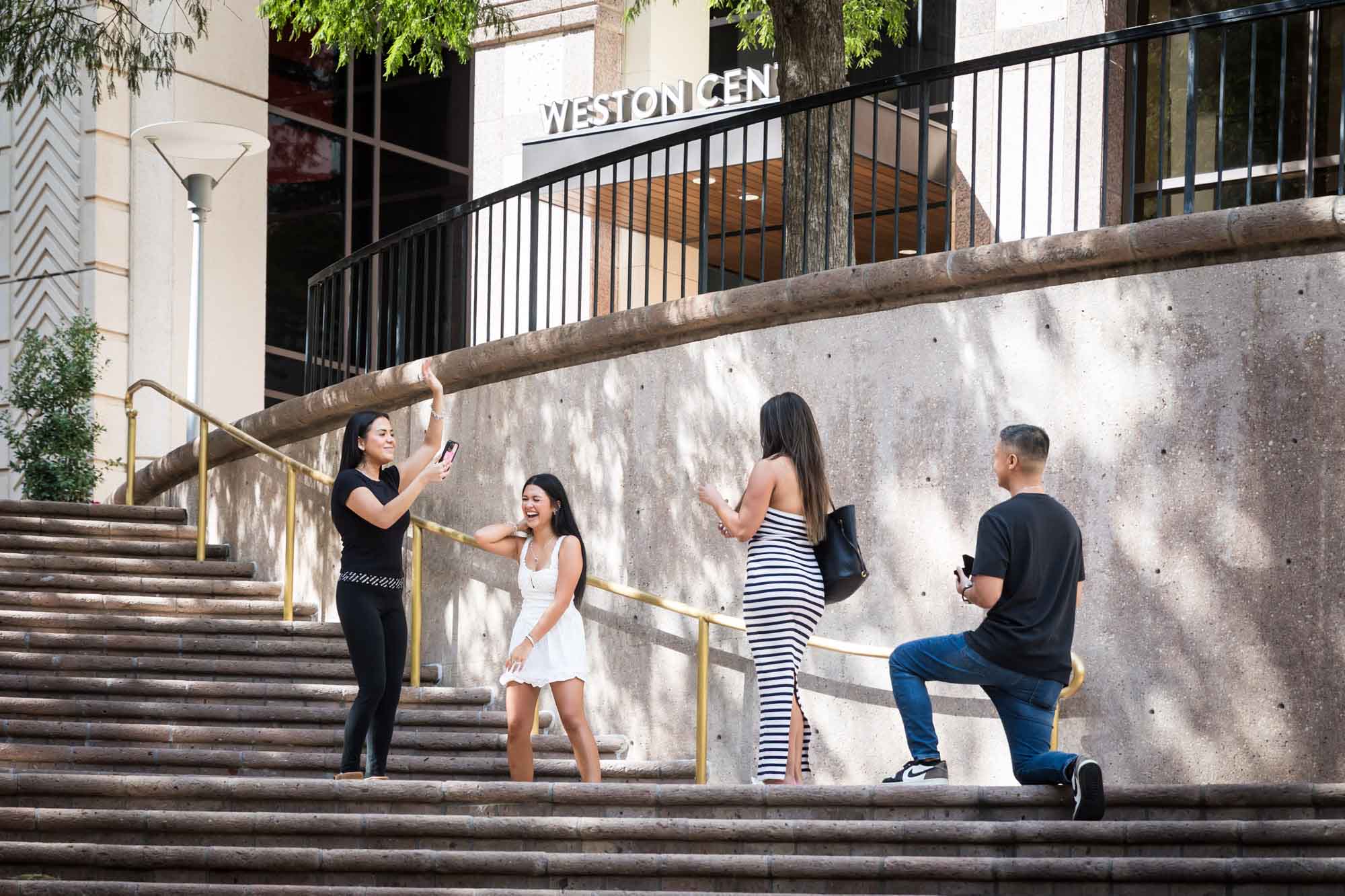Two young girls laughing and holding phone beside woman wearing black and white striped dress and man bent down on knee on stairs during Riverwalk surprise proposal in San Antonio