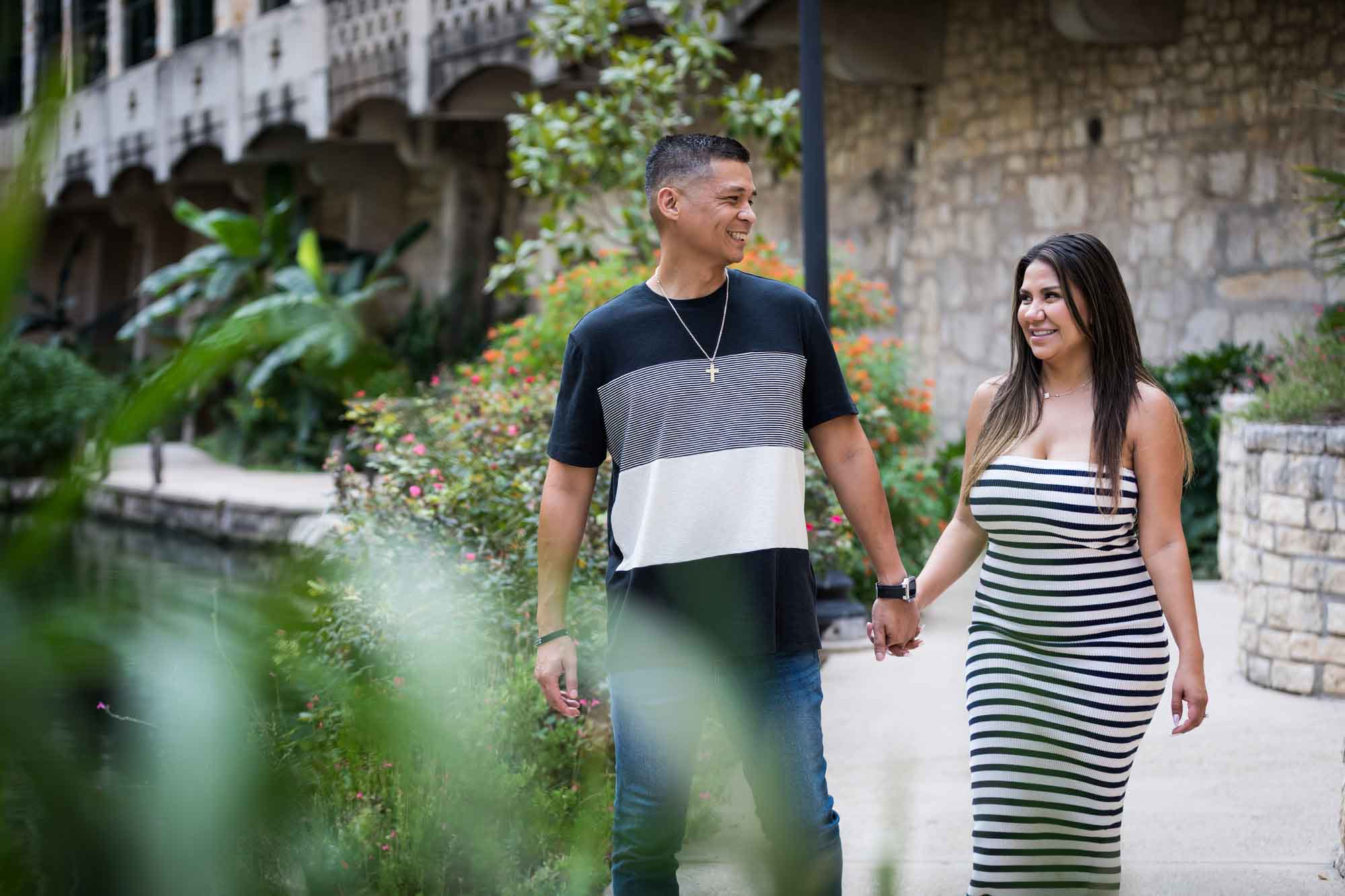 Man wearing striped shirt and jeans holding hands with woman wearing black and white striped, sleeveless dress during a Riverwalk engagement photo shoot