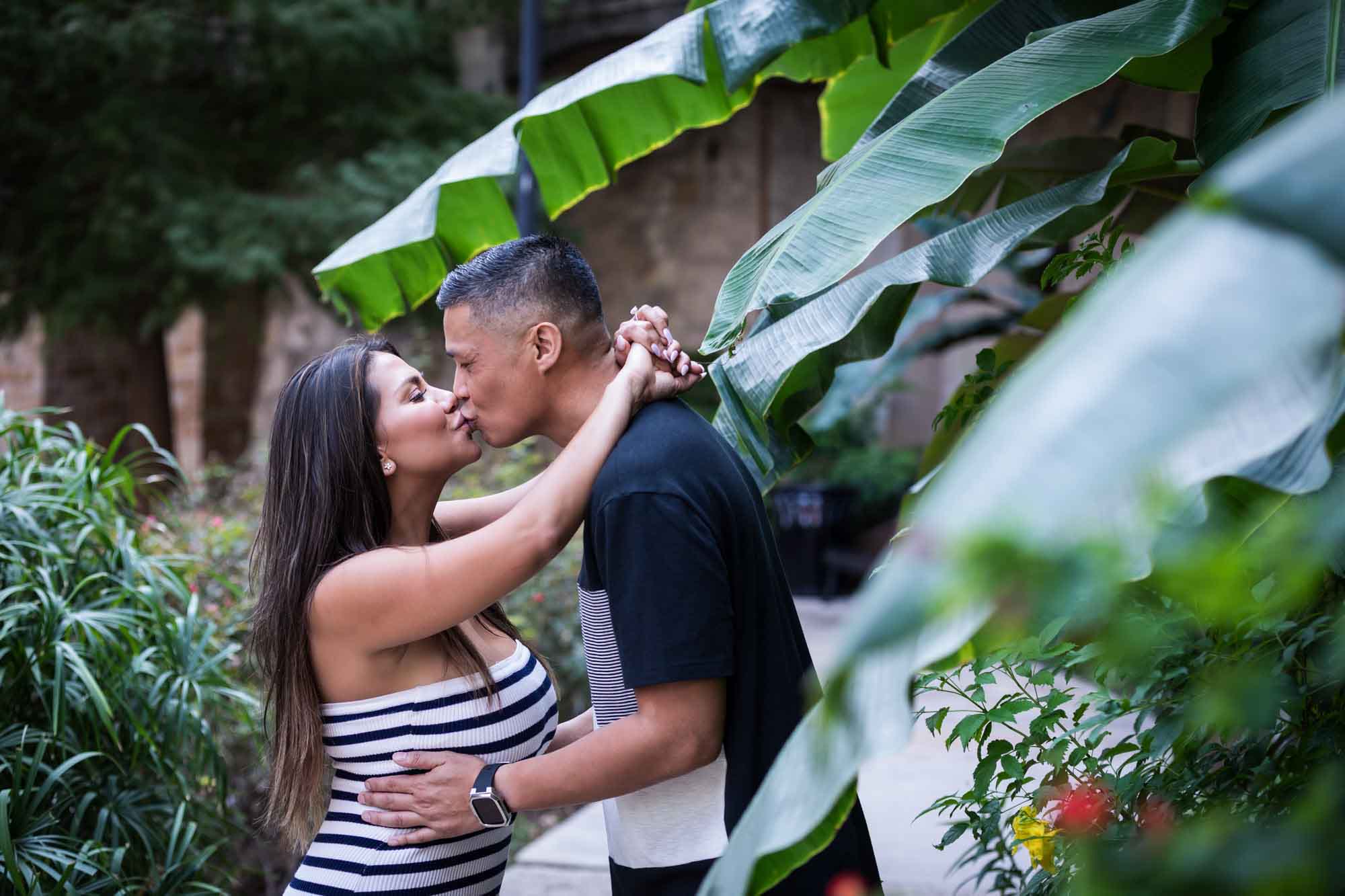 Man and woman kissing with woman's arms around man's neck and banana tree leaves in the background for an article entitled, ‘When Should I Book My Wedding Photographer?’