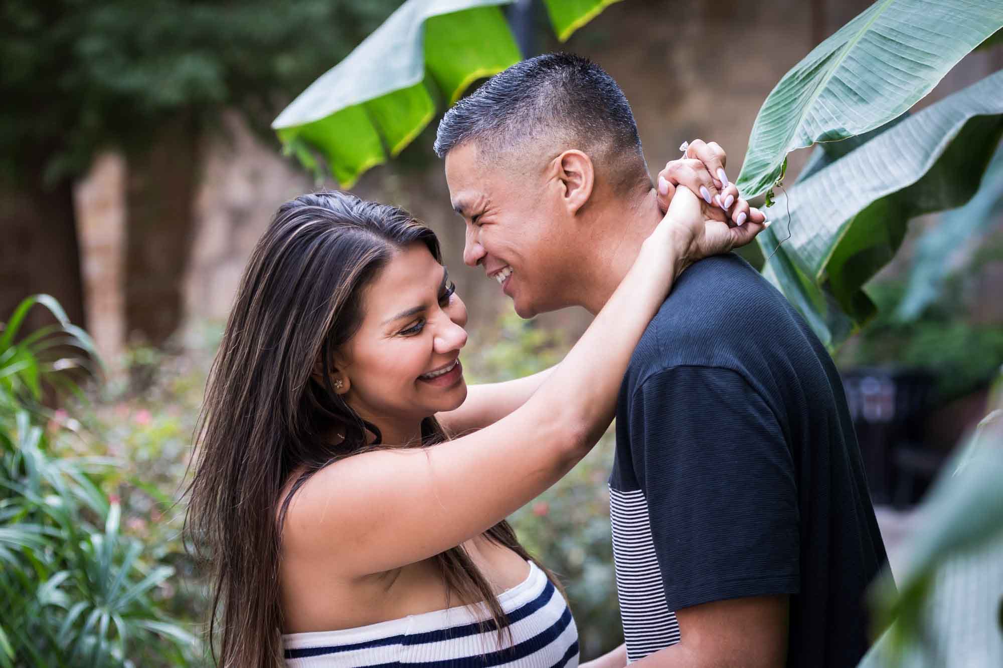 Man and woman laughing while woman's arms are around man's neck with banana tree leaves in the background for an article entitled, ‘When Should I Book My Wedding Photographer?’