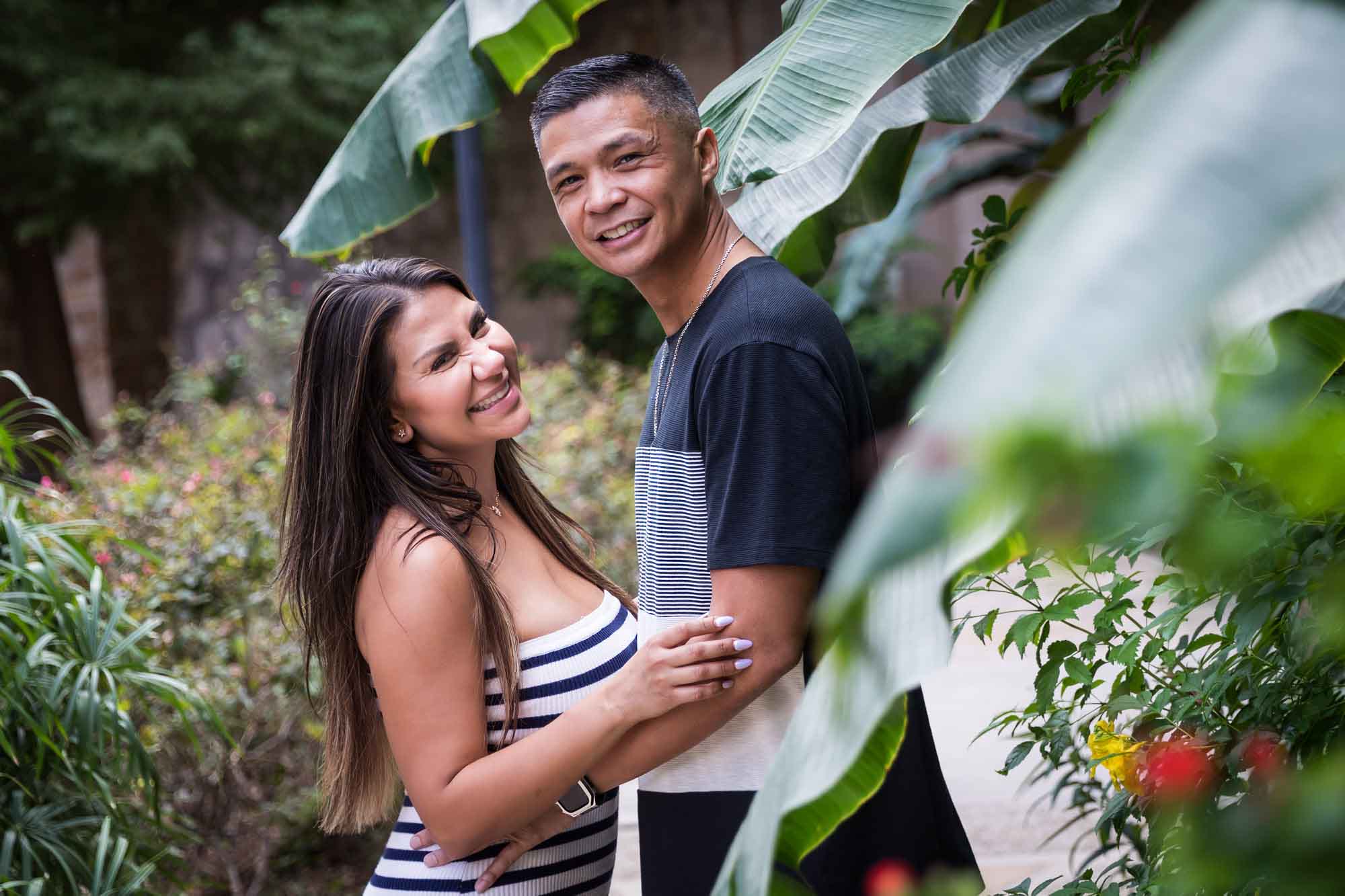 Man and woman hugging and touching arms while looking into camera behind banana tree leaves for an article entitled, ‘When Should I Book My Wedding Photographer?’