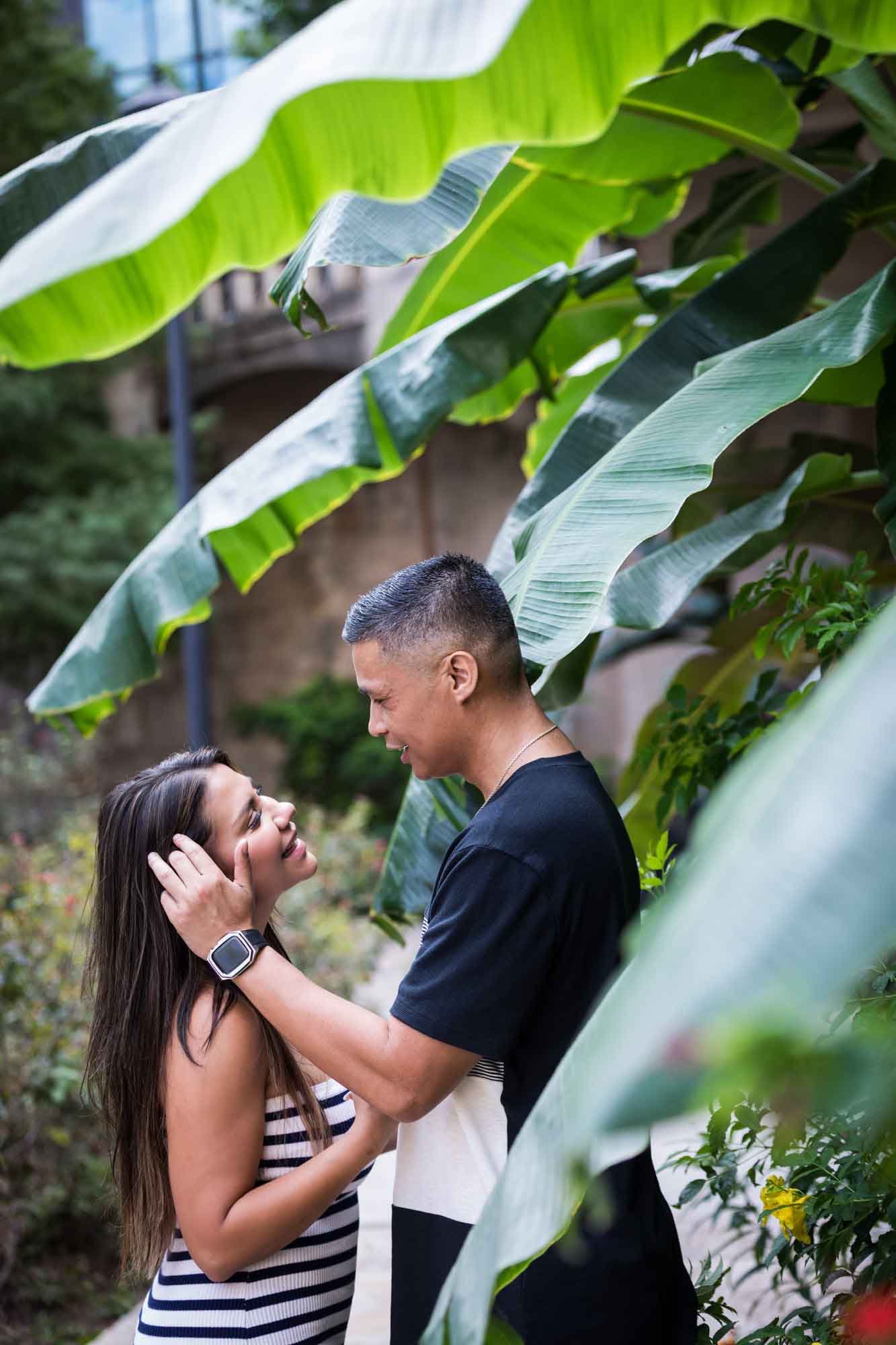 Man pushing hair back from woman's face standing in front of banana tree on Riverwalk for an article entitled, ‘When Should I Book My Wedding Photographer?’