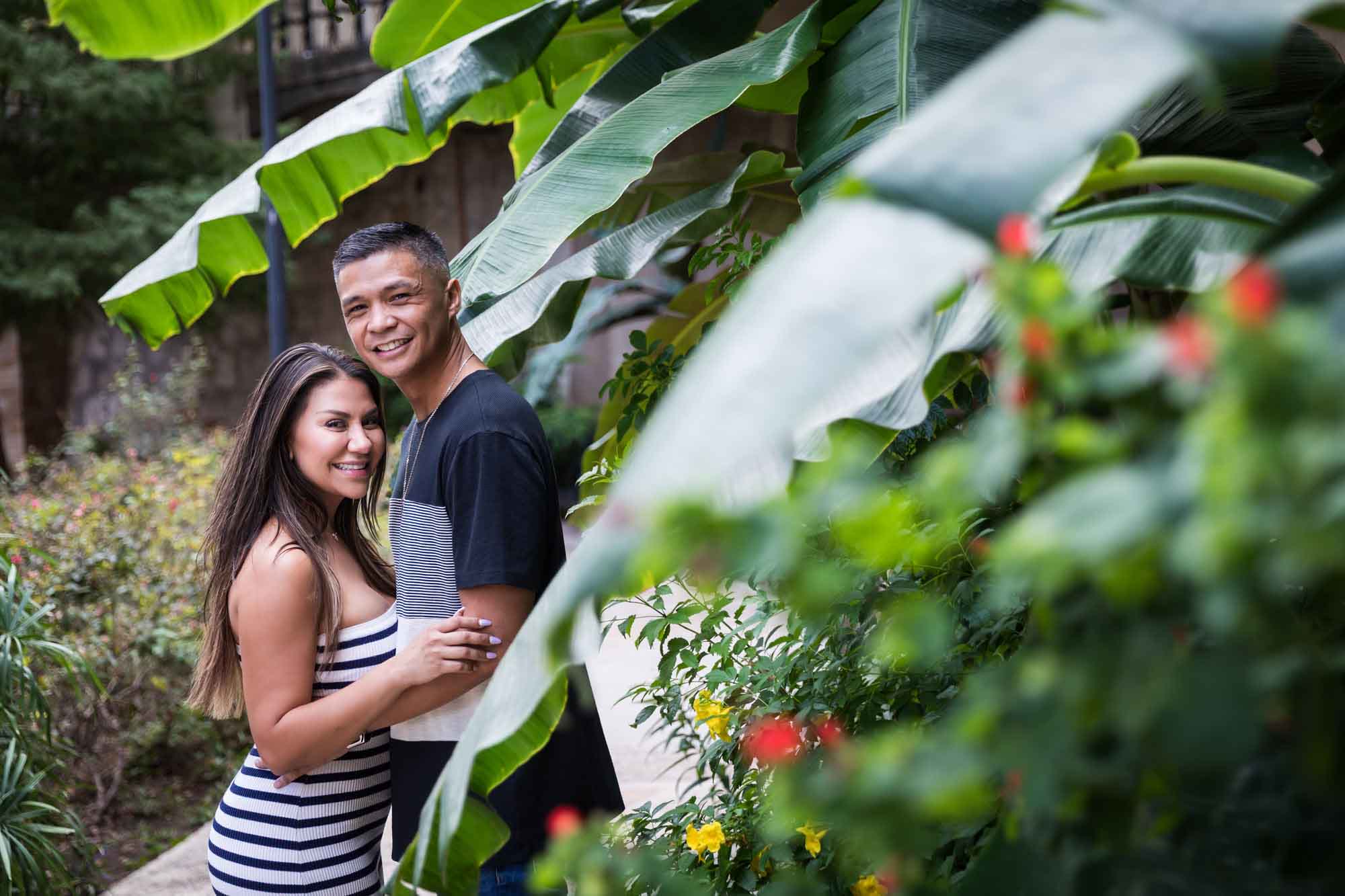 Man and woman hugging while looking into camera behind leaves of banana tree on Riverwalk for an article entitled, ‘When Should I Book My Wedding Photographer?’
