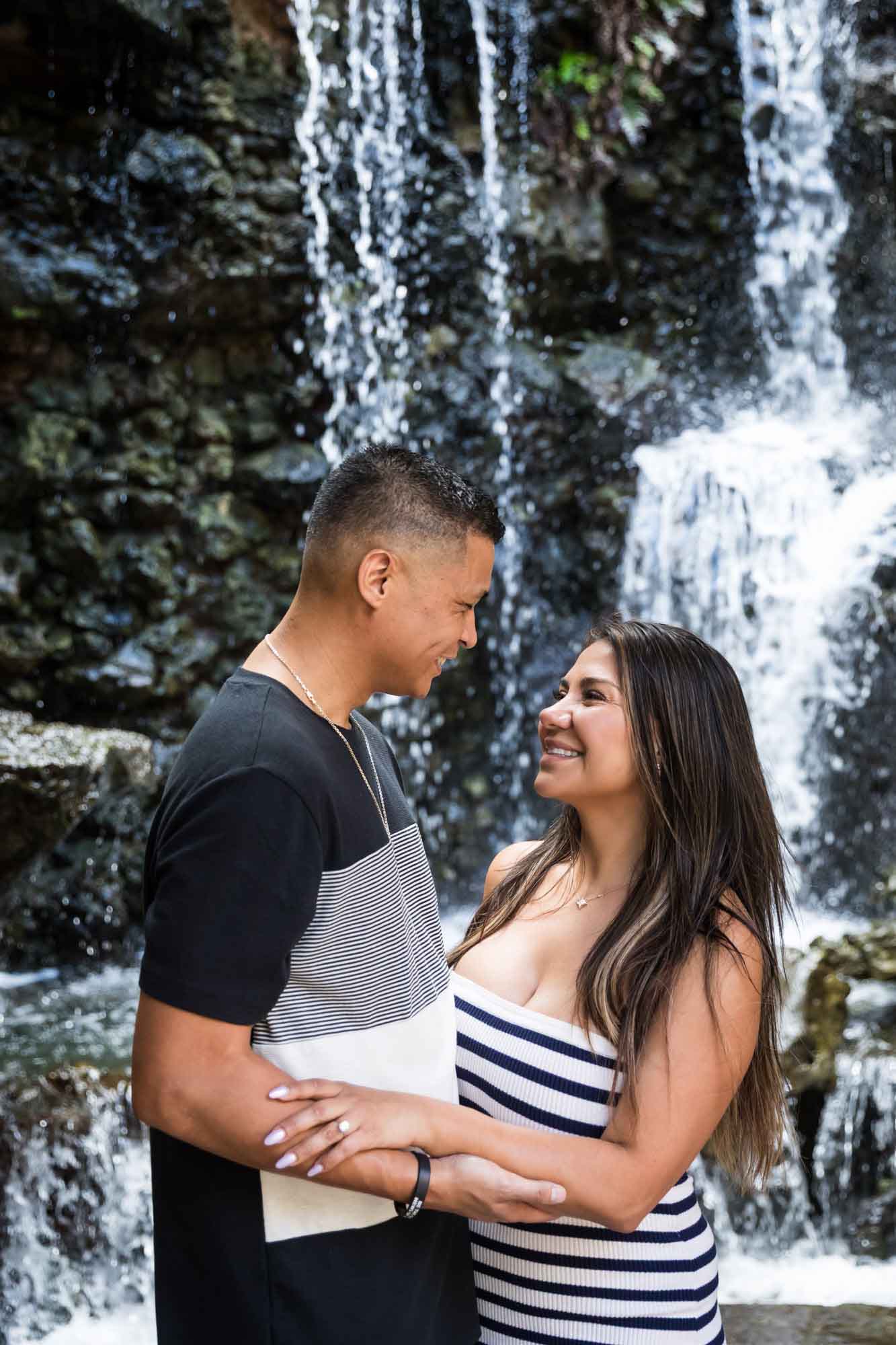 Man and woman looking at each other in front of waterfall while touching arms for an article entitled, ‘When Should I Book My Wedding Photographer?’