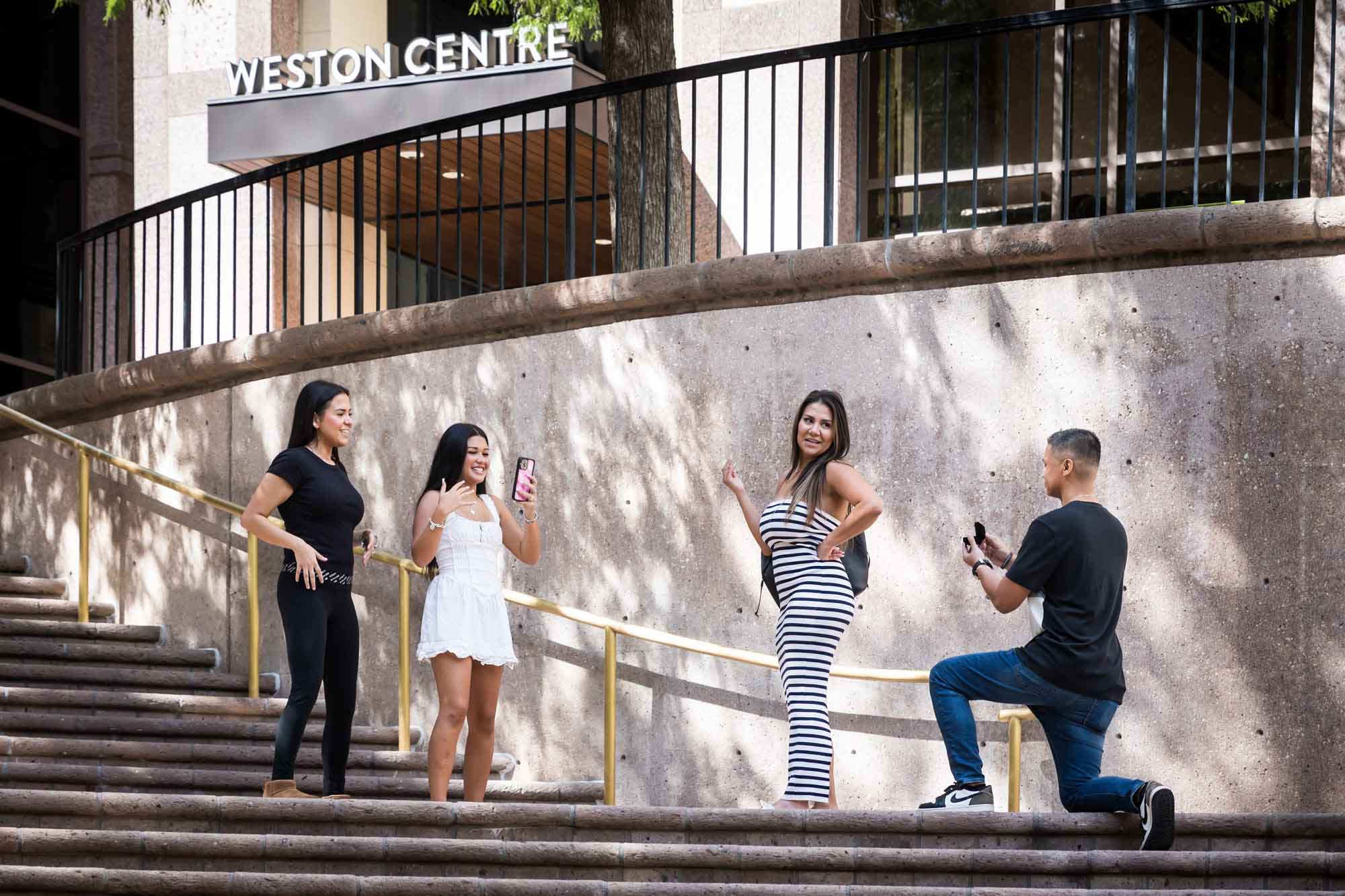 Two young girls laughing and holding phone beside woman wearing black and white striped dress smiling at man bent down on knee on stairs during Riverwalk surprise proposal in San Antonio