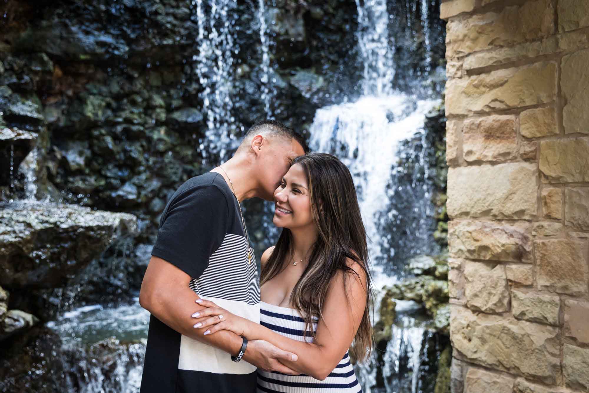 Man kissing woman on the side of the head in front of waterfall beside stone wall for an article entitled, ‘When Should I Book My Wedding Photographer?’