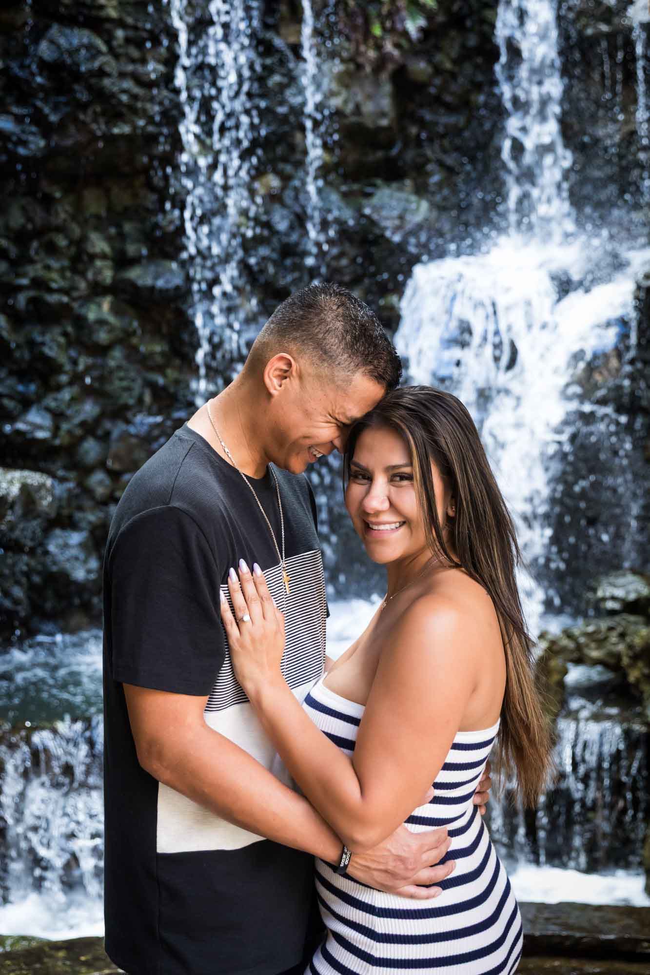 Man with forehead pressed against woman's head while she looks into camera in front of waterfall for an article entitled, ‘When Should I Book My Wedding Photographer?’