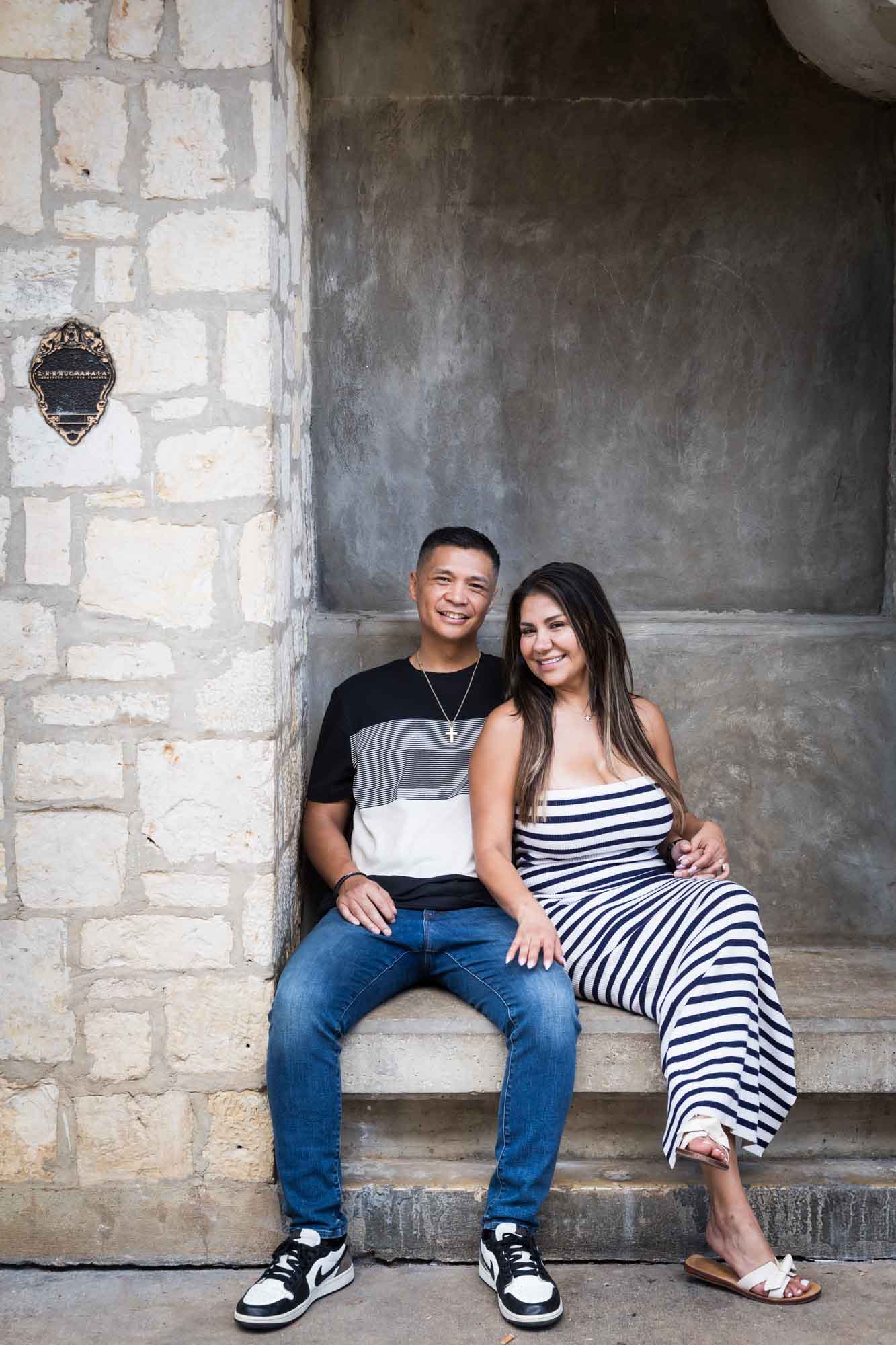 Couple sitting on stone bench beside stone wall with woman's hand on man's knee for an article entitled, ‘When Should I Book My Wedding Photographer?’