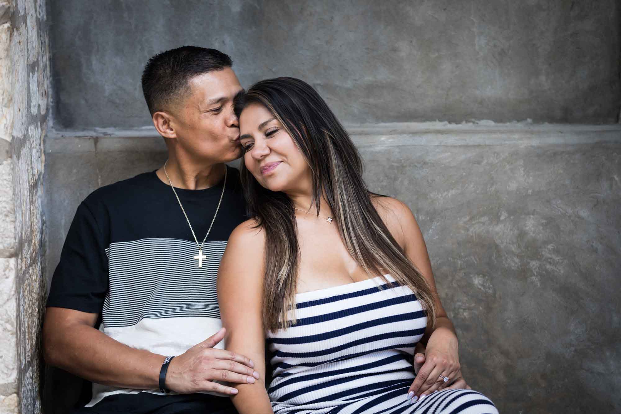 Man kissing woman on the side of the head in front of stone wall for an article entitled, ‘When Should I Book My Wedding Photographer?’