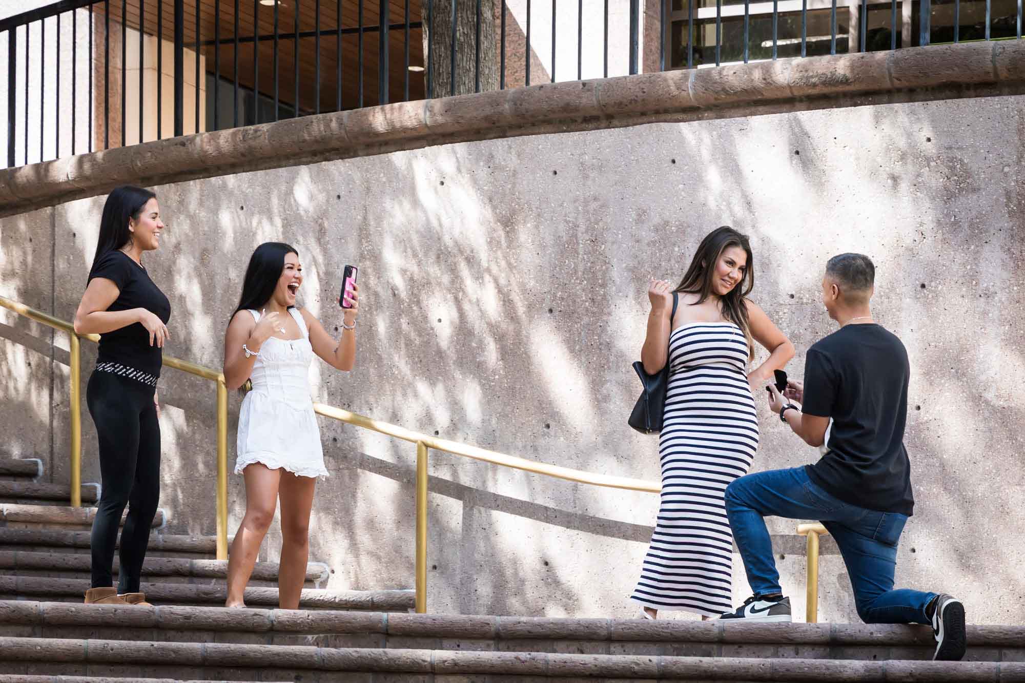 Two young girls laughing and holding phone beside woman wearing black and white striped dress smiling at man bent down on knee on stairs during Riverwalk surprise proposal in San Antonio