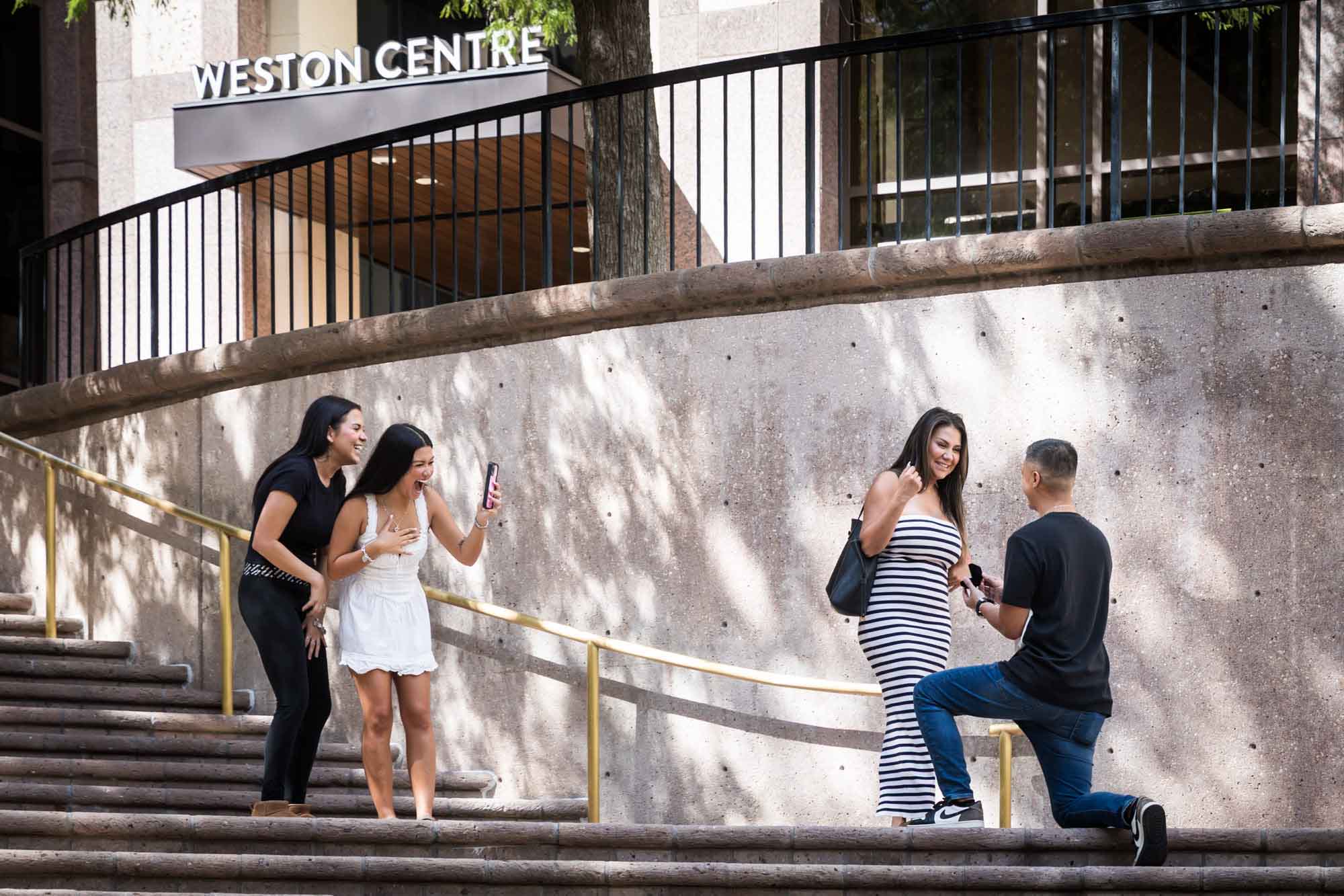 Two young girls laughing and holding phone beside woman wearing black and white striped dress smiling at man bent down on knee on stairs during Riverwalk surprise proposal in San Antonio
