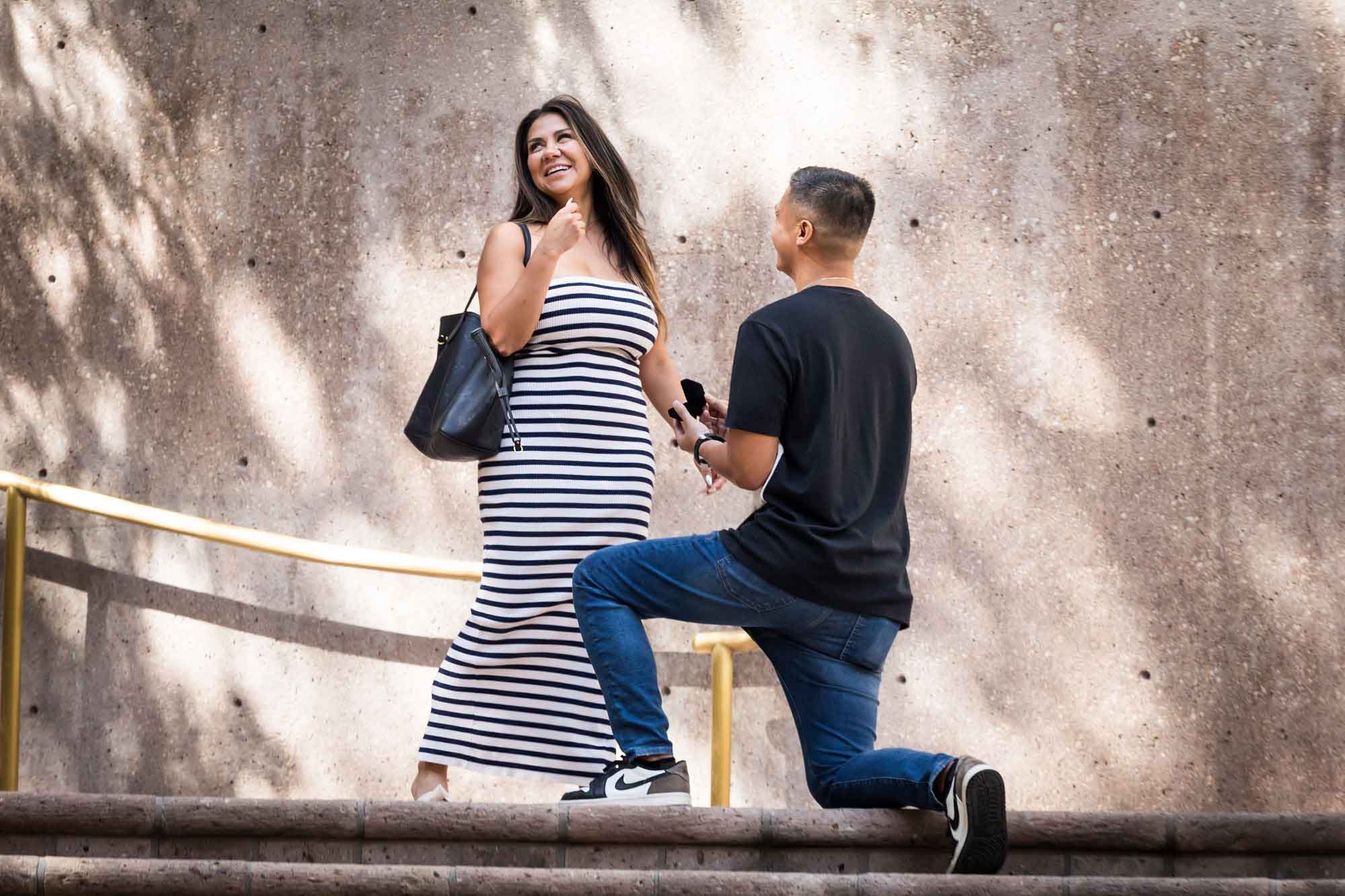 Woman wearing black and white striped dress smiling in front of man bent down on knee on stairs during Riverwalk surprise proposal in San Antonio