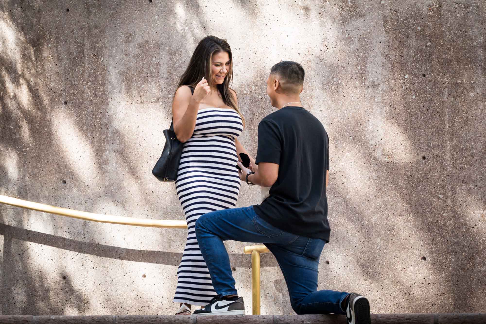 Woman wearing black and white striped dress smiling at man bent down on knee on stairs during Riverwalk surprise proposal in San Antonio
