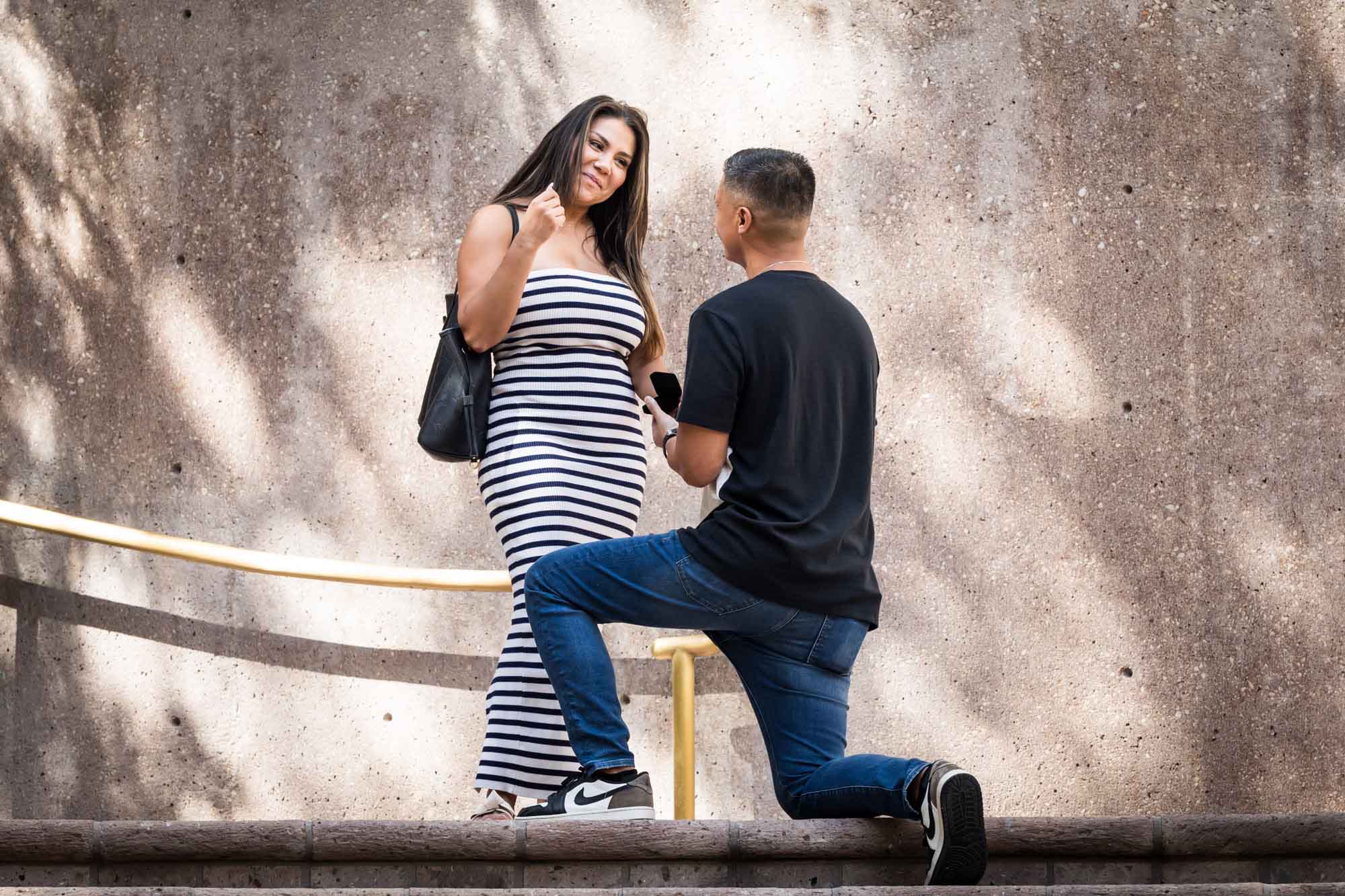 Woman wearing black and white striped dress smiling at man bent down on knee on stairs during Riverwalk surprise proposal in San Antonio