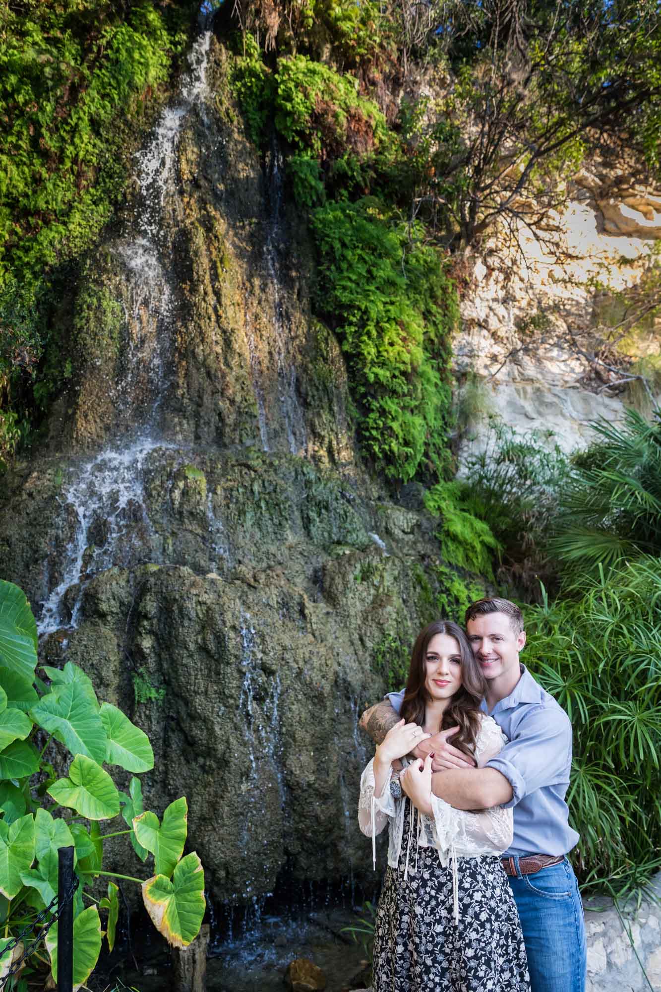 Couple hugging in front of moss-covered waterfall during a Japanese Tea Garden engagement portrait 