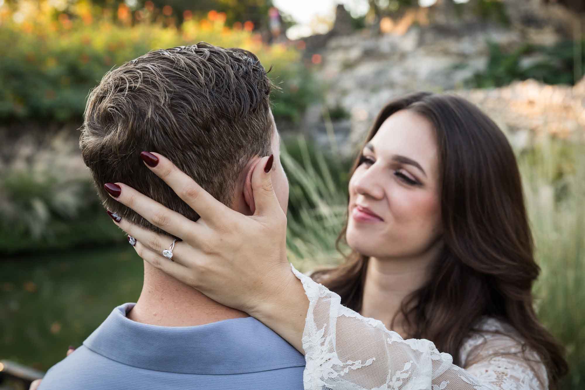 Close up of a woman's hand wearing an engagement ring on the back of a man's neck as he looks at a woman for an article on ‘Where do I Start with Wedding Planning?’