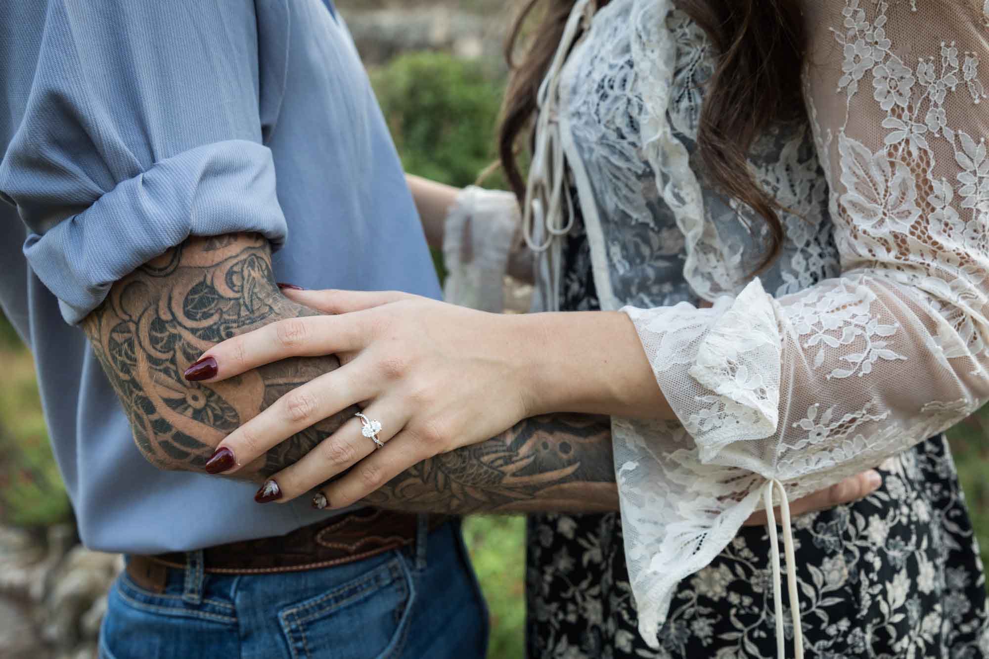 Close up of a woman's hand wearing an engagement ring resting on a man's arm with tattoos for an article on ‘Where do I Start with Wedding Planning?’