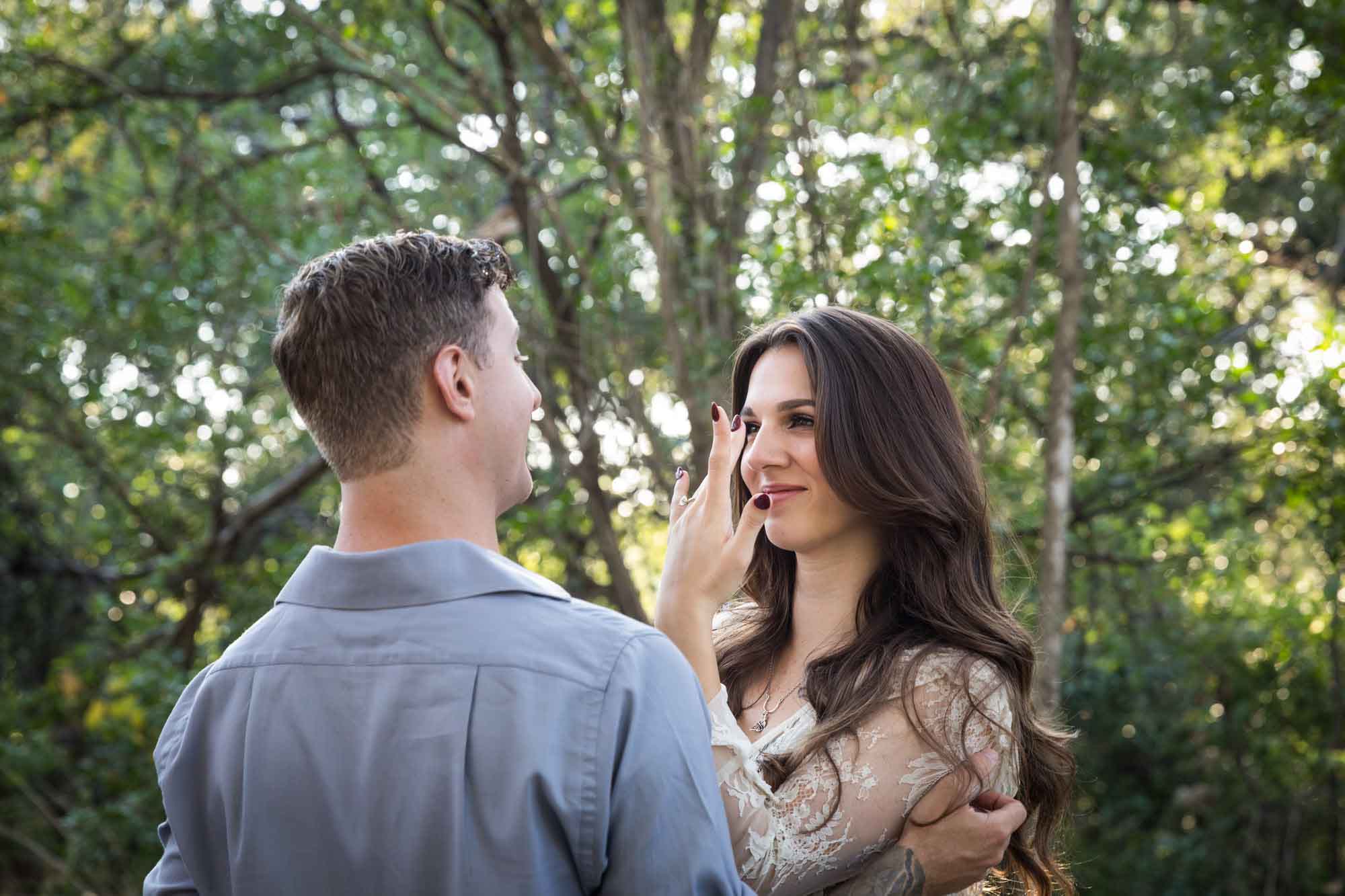 Woman showing hand to man under tree branches in Brackenridge Park during a Japanese Tea Garden engagement portrait by San Antonio wedding photographer Kelly Williams