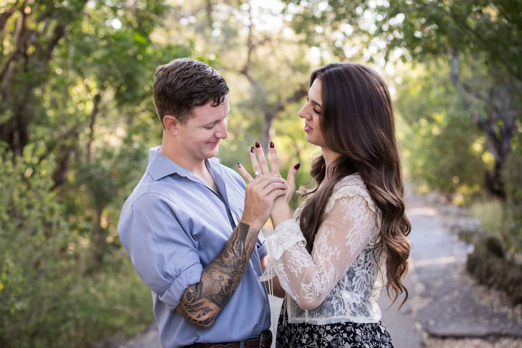Couple looking at engagement ring under tree branches in Brackenridge Park during a Japanese Tea Garden engagement portrait by San Antonio wedding photographer Kelly Williams