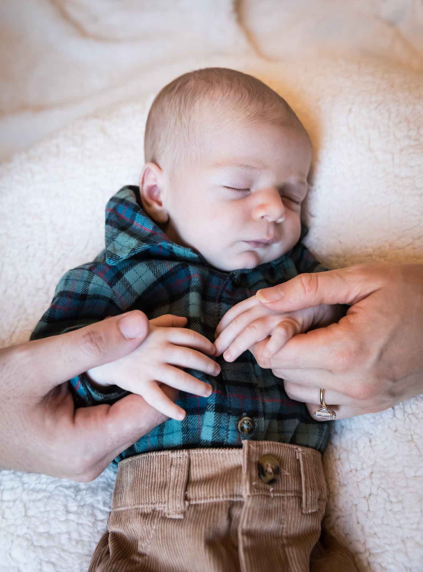 Hands of mother and father holding hands of baby boy wearing green and blue plaid shirt