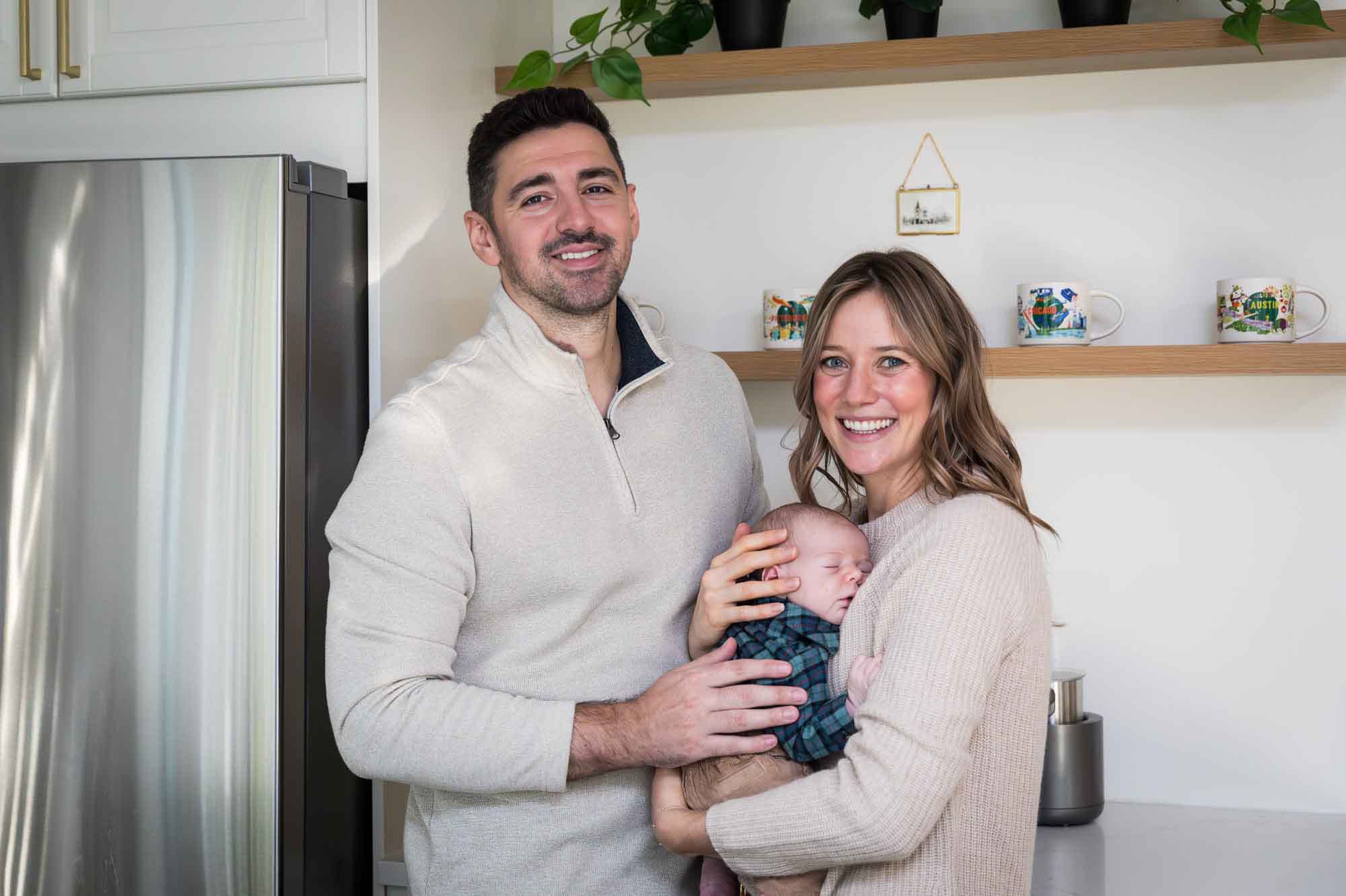 Mother and father holding baby boy wearing plaid shirt standing in white kitchen during an Austin newborn portrait session