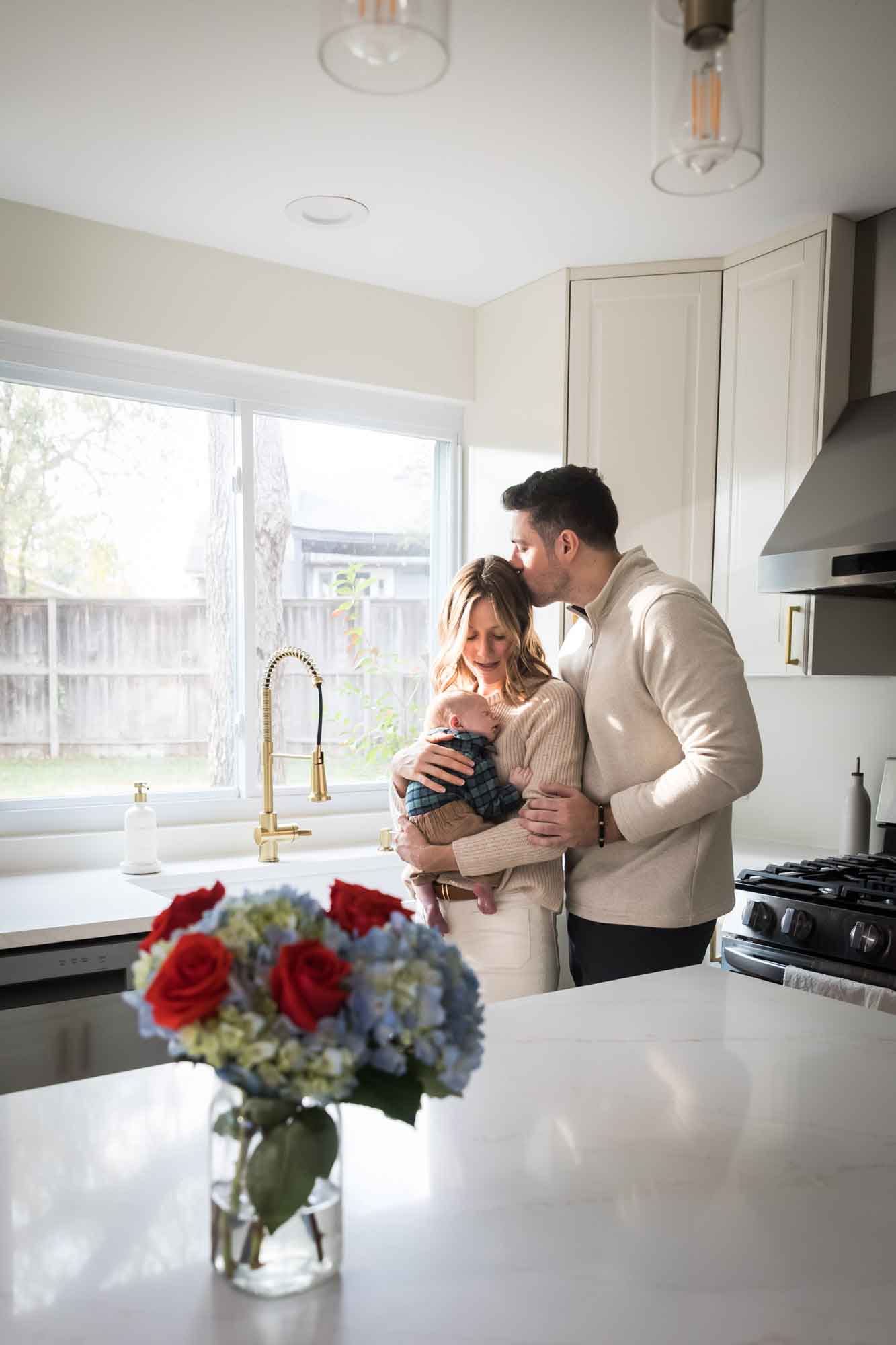 Mother holding baby boy wearing plaid shirt and father kissing mother while standing in white kitchen during an Austin newborn portrait session