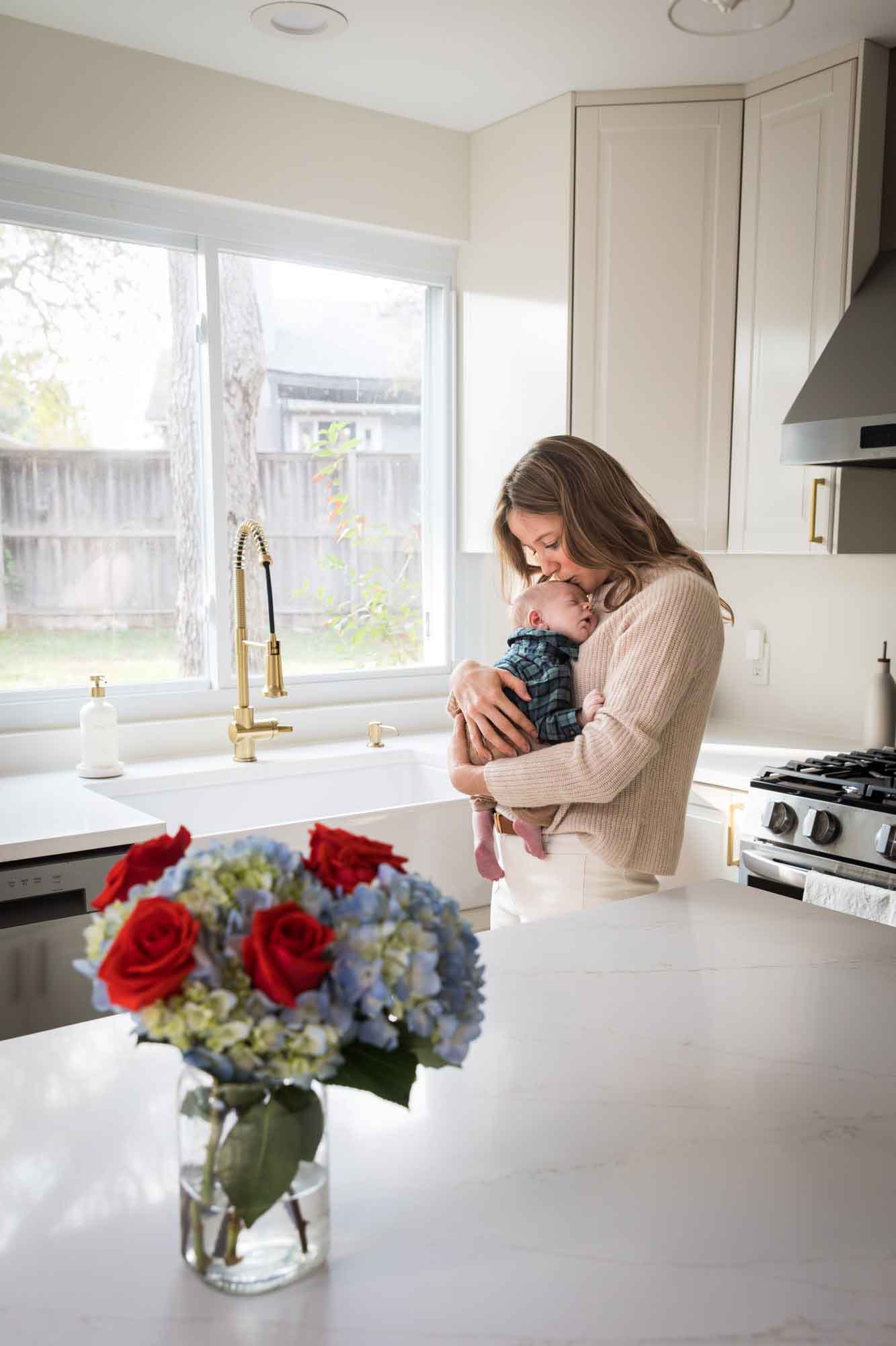 Mother kissing baby boy wearing plaid shirt in front of white kitchen sink and cabinets during an Austin newborn portrait session