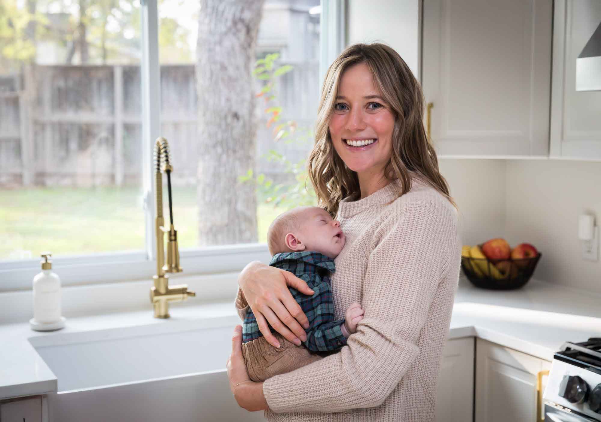 Mother holding baby boy wearing plaid shirt in front of white kitchen sink and cabinets during an Austin newborn portrait session