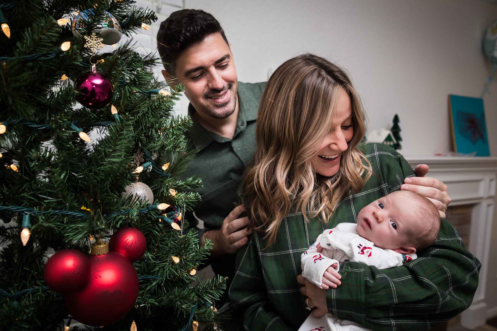 Mother and father looking down at newborn baby boy in front of Christmas tree during an Austin newborn portrait session