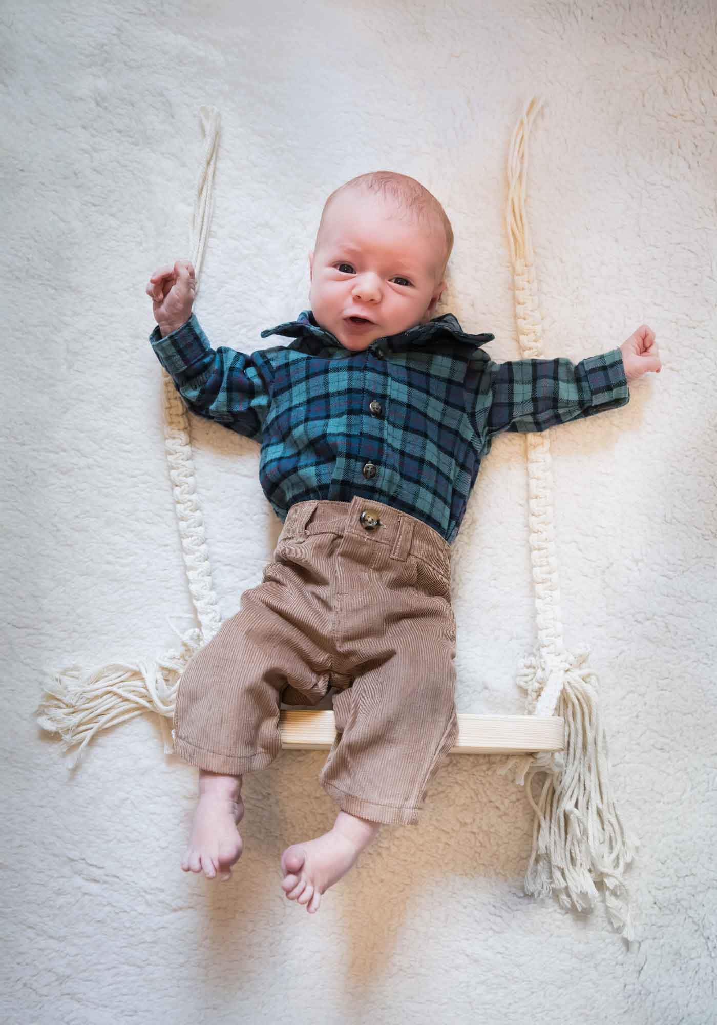 Baby boy wearing green and blue plaid shirt and brown corduroy pants resting on wooden swing on white, fleece blanket during an Austin newborn portrait session