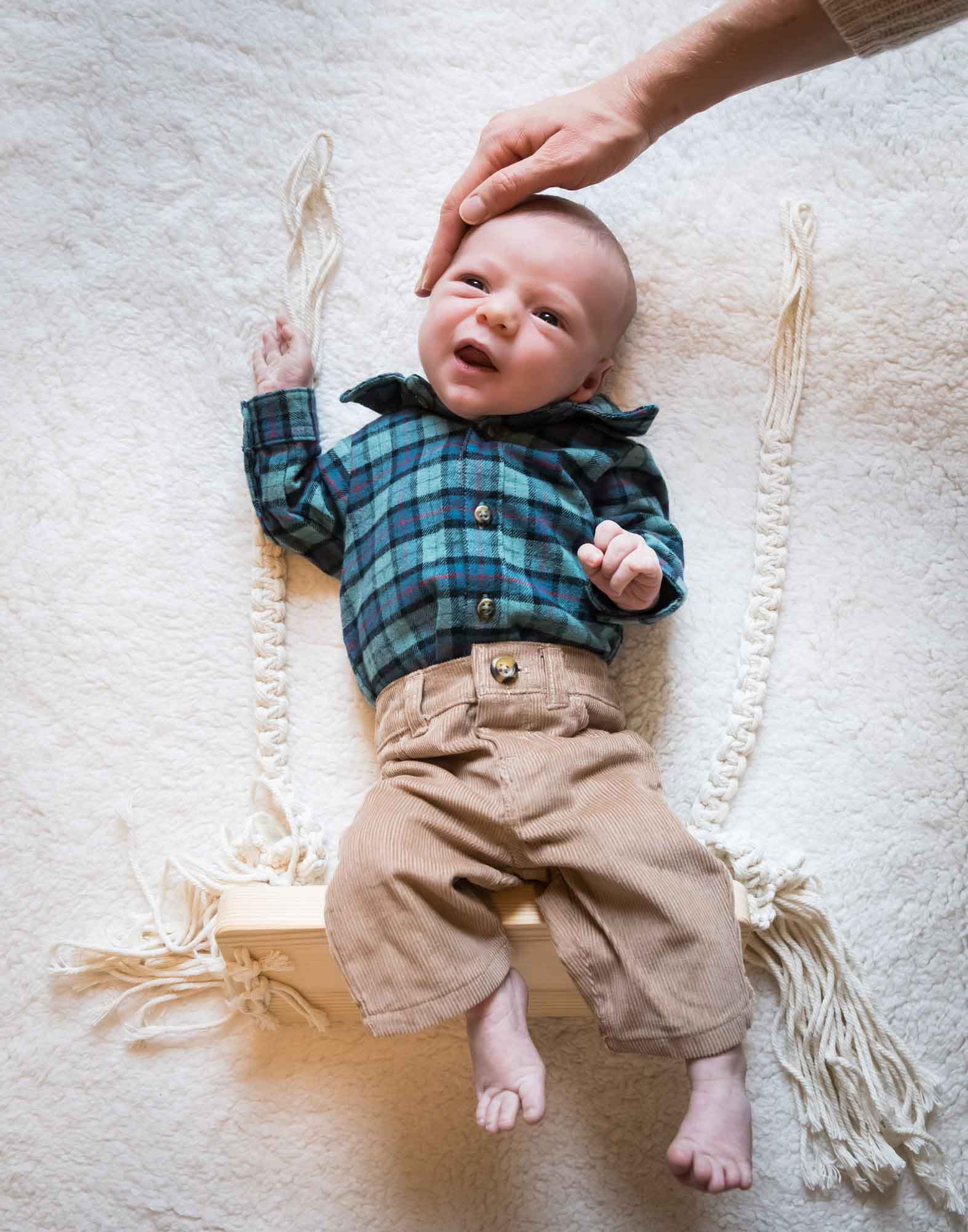 Baby boy wearing green and blue plaid shirt and brown corduroy pants resting on wooden swing on white, fleece blanket during an Austin newborn portrait session