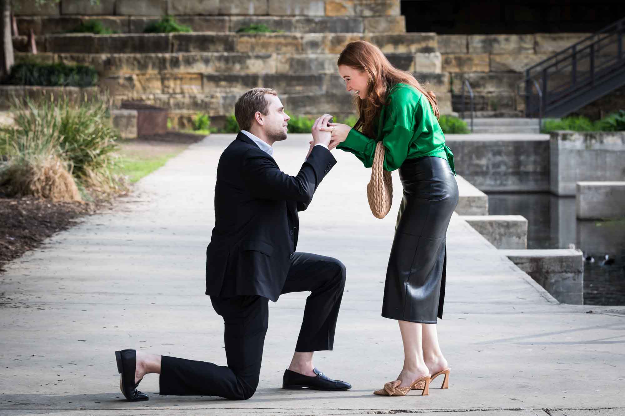 Man wearing black suit proposing while down on one knee in front of woman wearing green blouse on sidewalk in front of Emma Hotel beside Riverwalk during an Emma Hotel surprise proposal