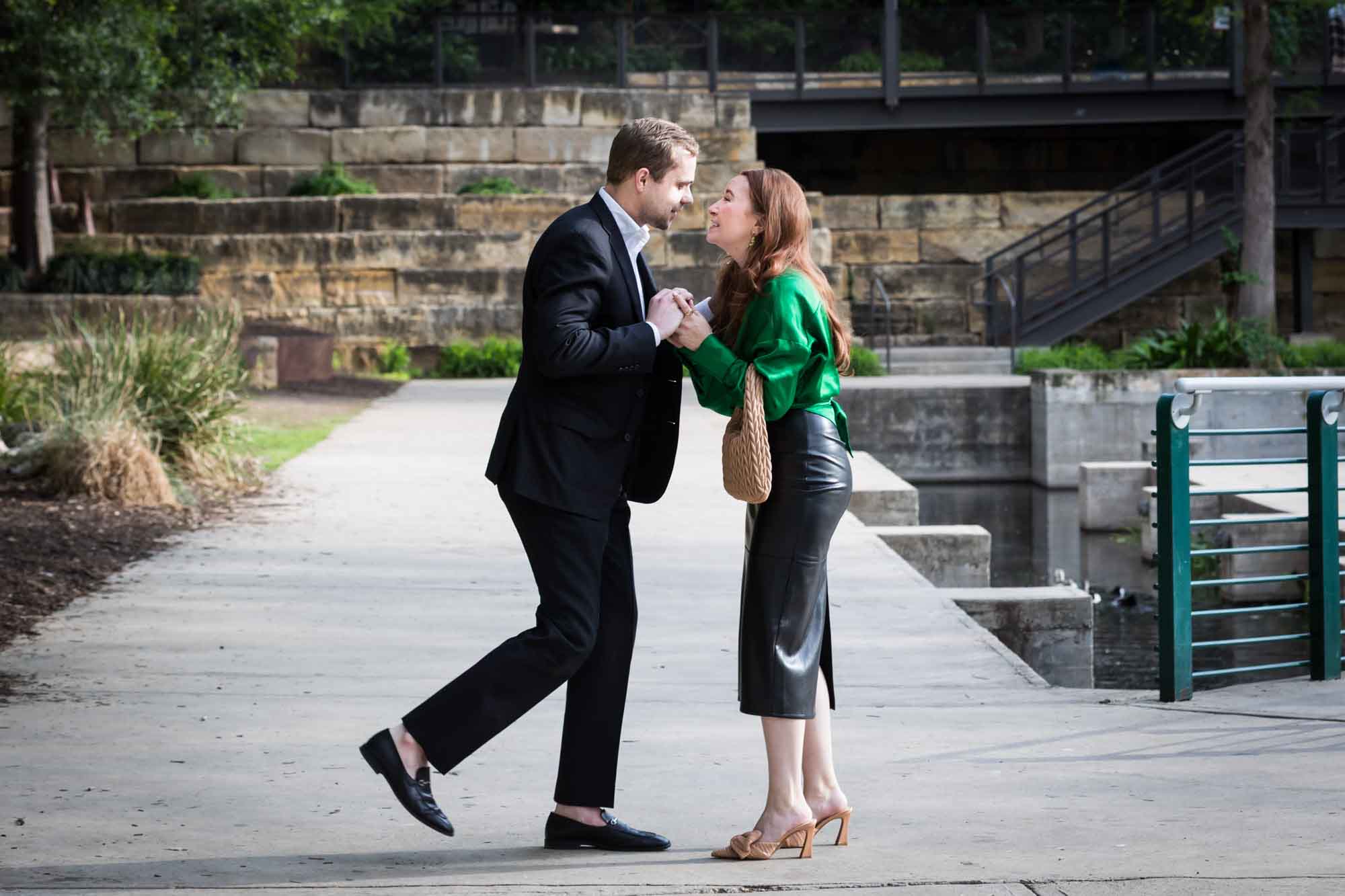 Man wearing black suit standing in front of woman wearing green blouse on sidewalk in front of Emma Hotel beside Riverwalk during an Emma Hotel surprise proposal