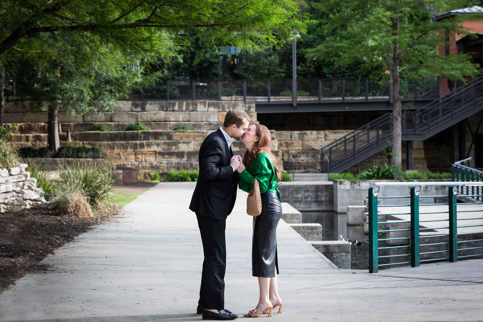 Man wearing black suit kissing woman wearing green blouse on sidewalk in front of Emma Hotel beside Riverwalk during an Emma Hotel surprise proposal