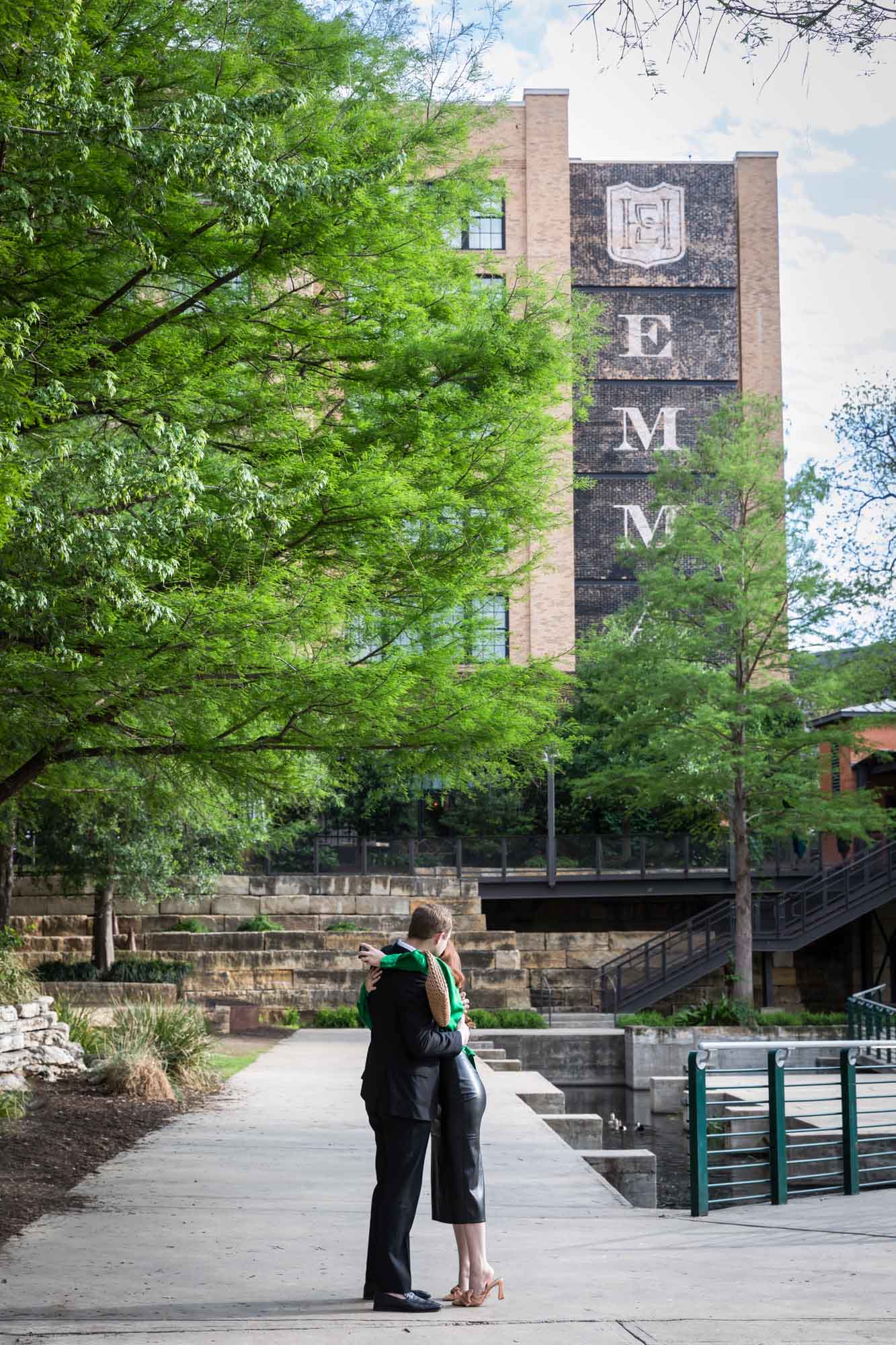 Man wearing black suit hugging woman wearing green blouse on sidewalk in front of Emma Hotel beside Riverwalk during an Emma Hotel surprise proposal
