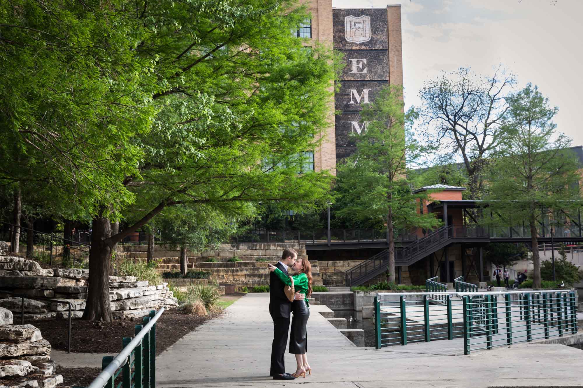Man wearing black suit kissing woman wearing green blouse on sidewalk in front of Emma Hotel beside Riverwalk during an Emma Hotel surprise proposal