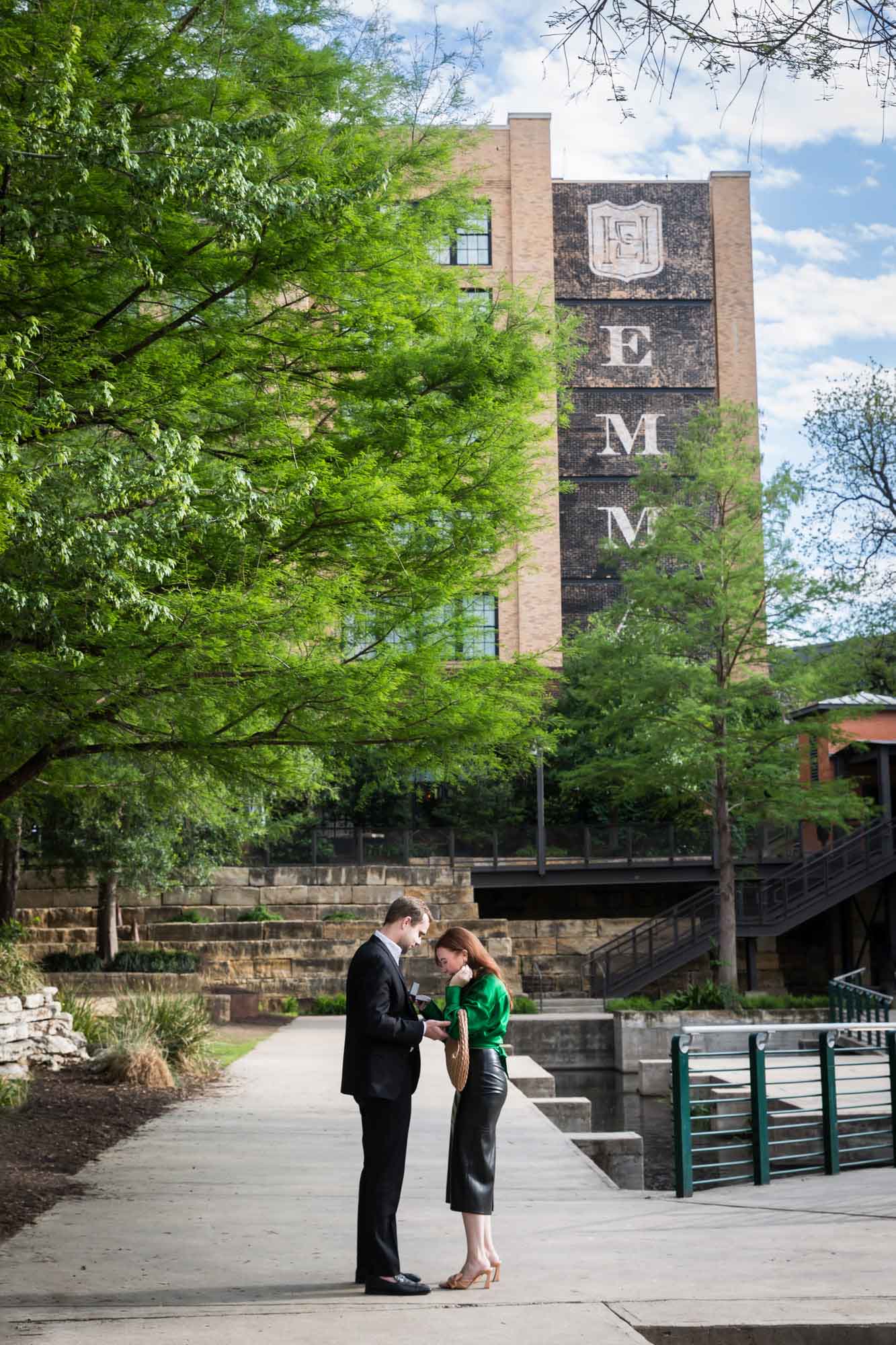Man wearing black suit standing in front of woman wearing green blouse on sidewalk in front of Emma Hotel beside Riverwalk during an Emma Hotel surprise proposal