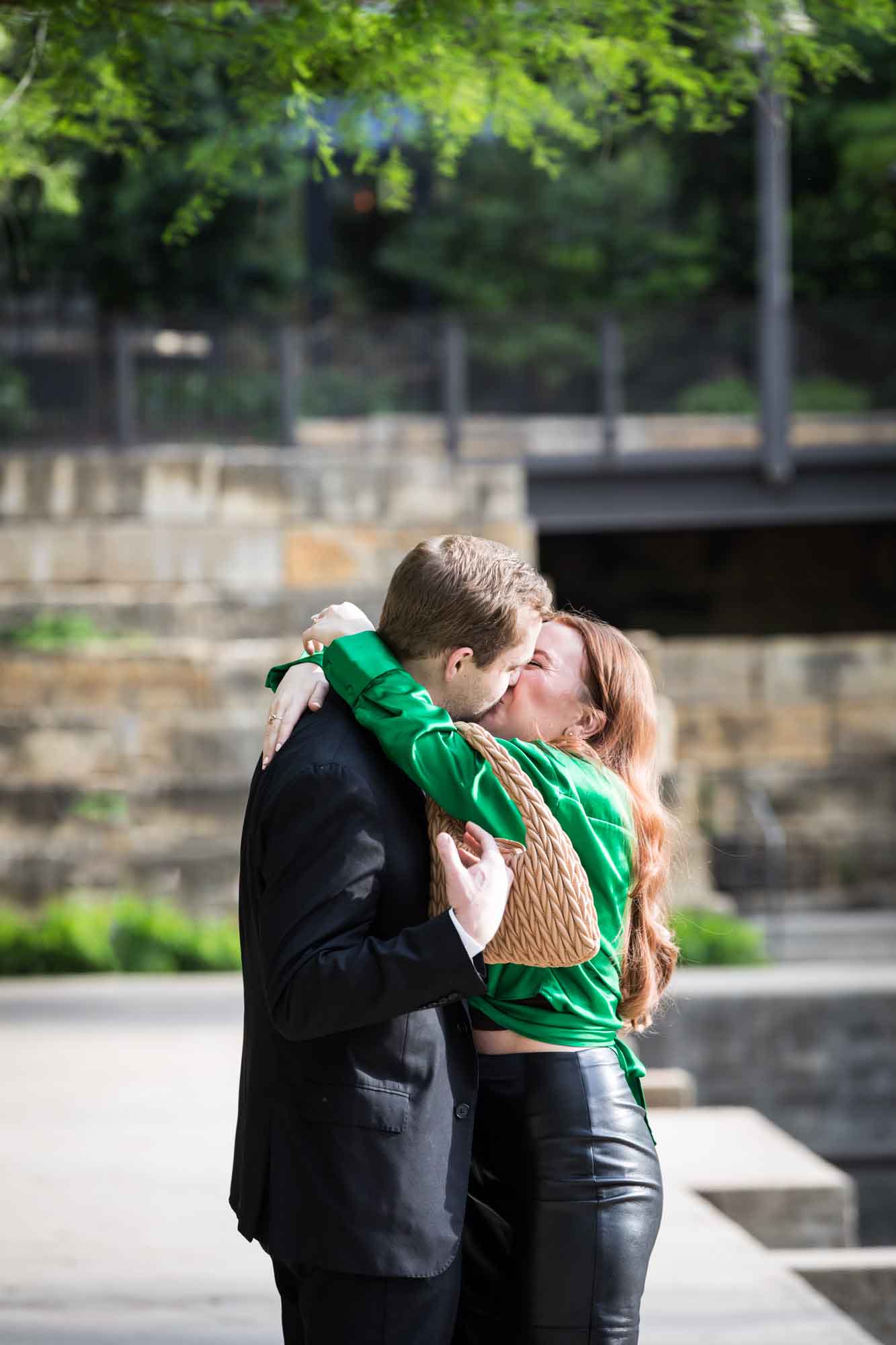 Man wearing black suit kissing woman wearing green blouse on sidewalk in front of Emma Hotel beside Riverwalk during an Emma Hotel surprise proposal