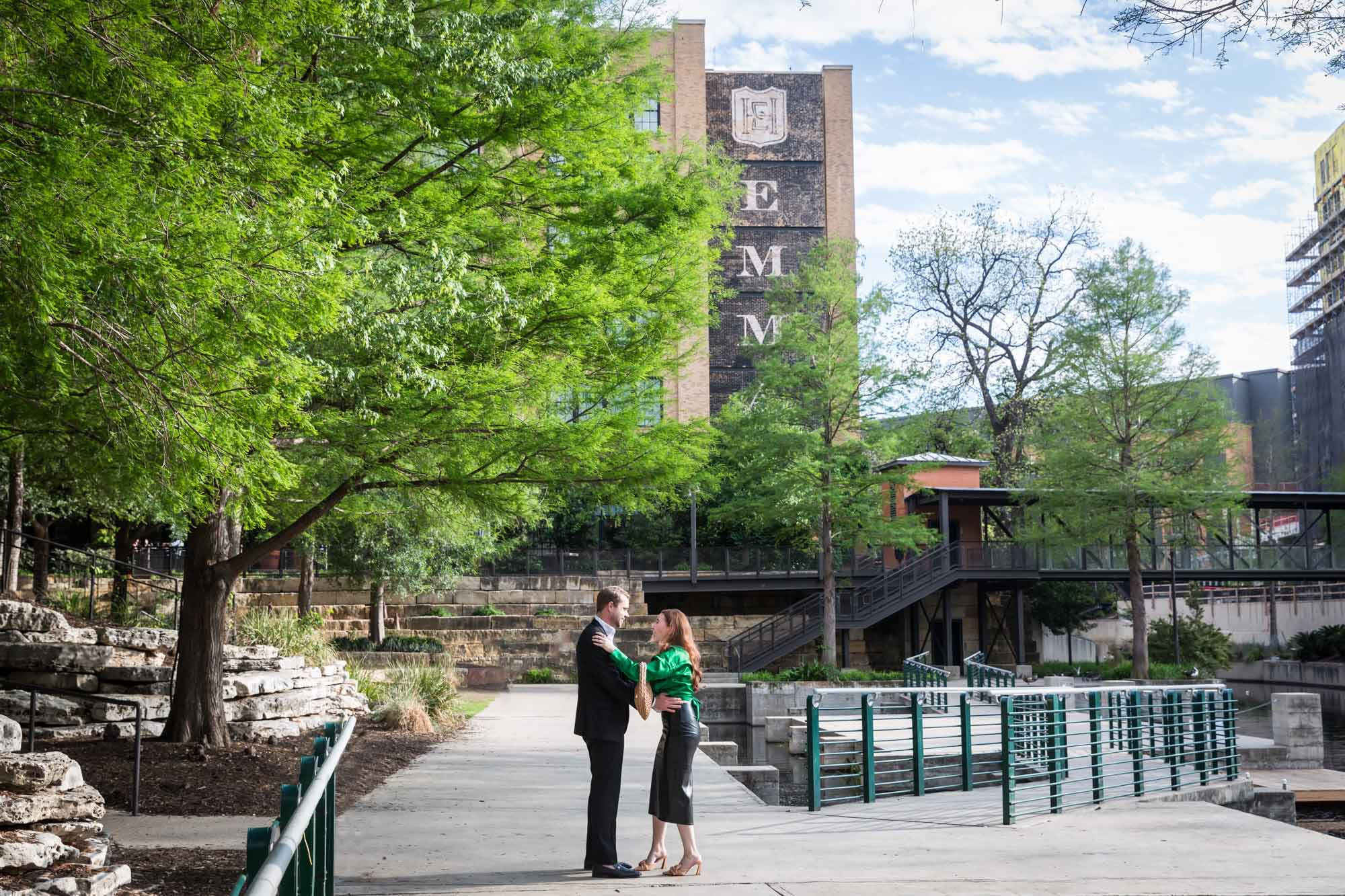 Man wearing black suit standing in front of woman wearing green blouse on sidewalk in front of Emma Hotel beside Riverwalk during an Emma Hotel surprise proposal