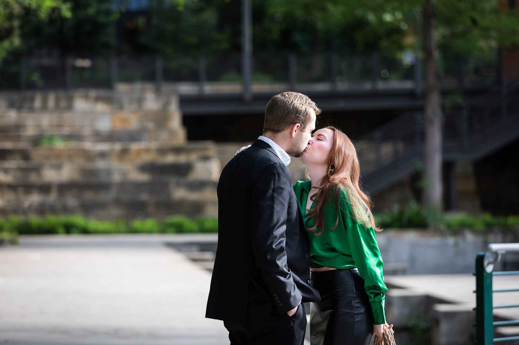 Man wearing black suit kissing woman wearing green blouse on sidewalk in front of Emma Hotel beside Riverwalk during an Emma Hotel surprise proposal